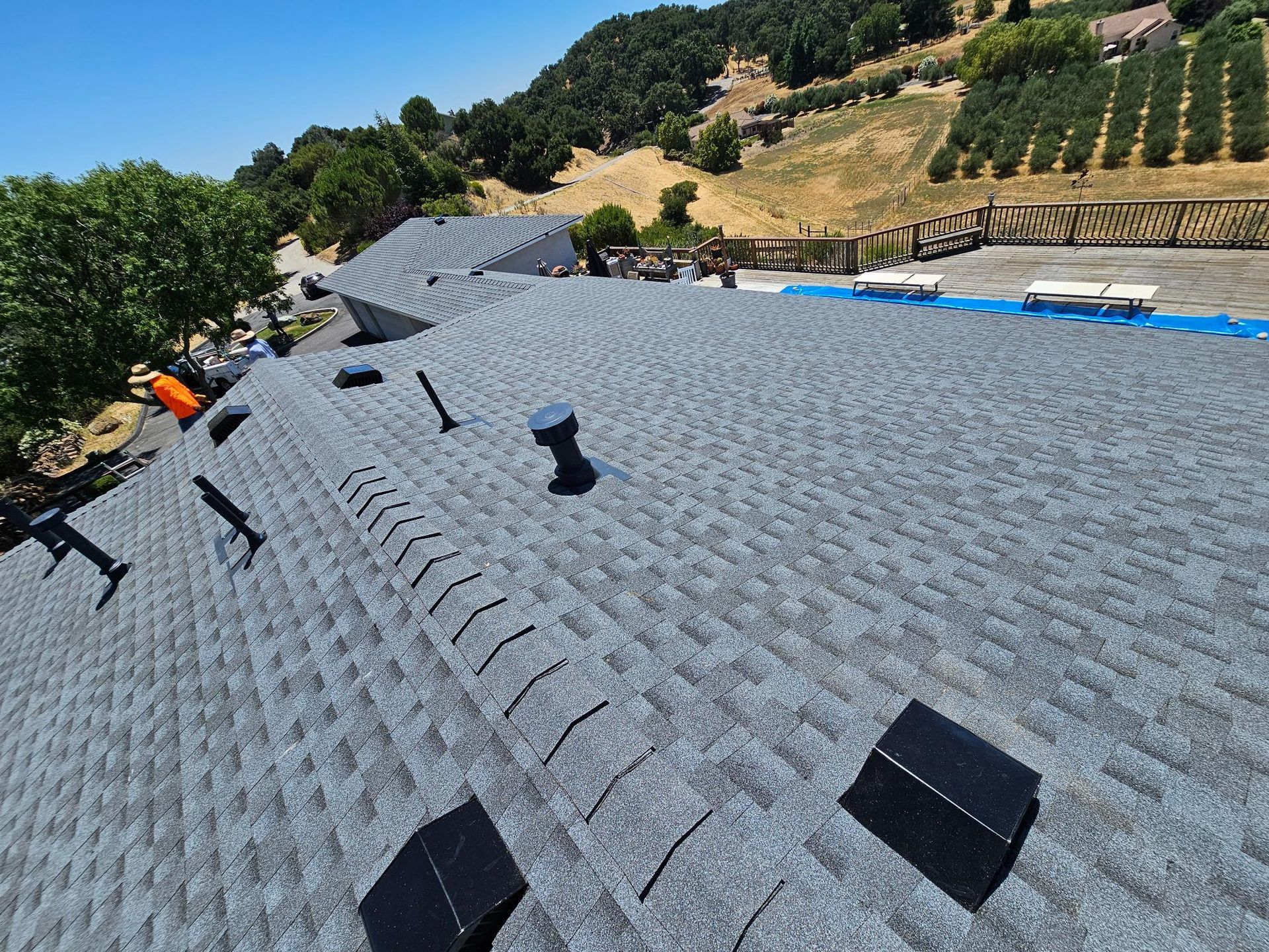 Gray shingled roof under construction with vents, blue tarp, and trees on a hillside in the background