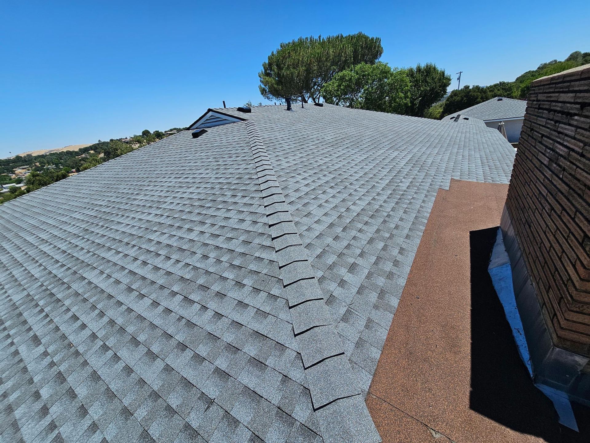 Gray shingle roof with roof vent and trees under a clear blue sky