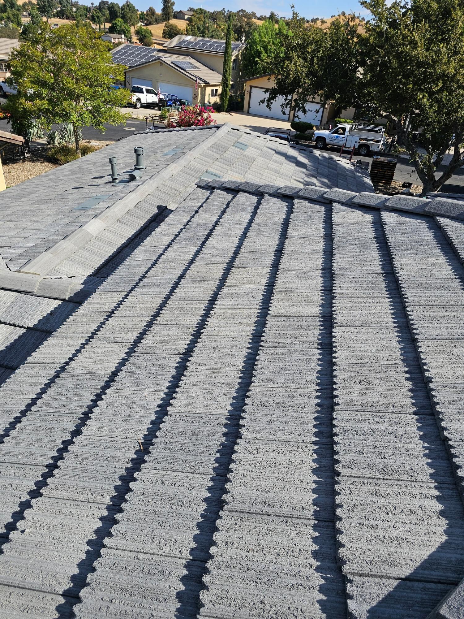 Roof covered in gray shingles with long dark shadows, viewed from the peak toward houses and trees below