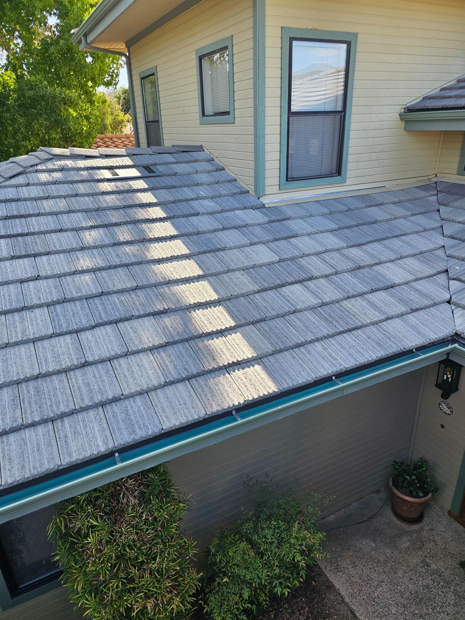 Slate-shingled roof with skylight beside a beige house and shrubs below.