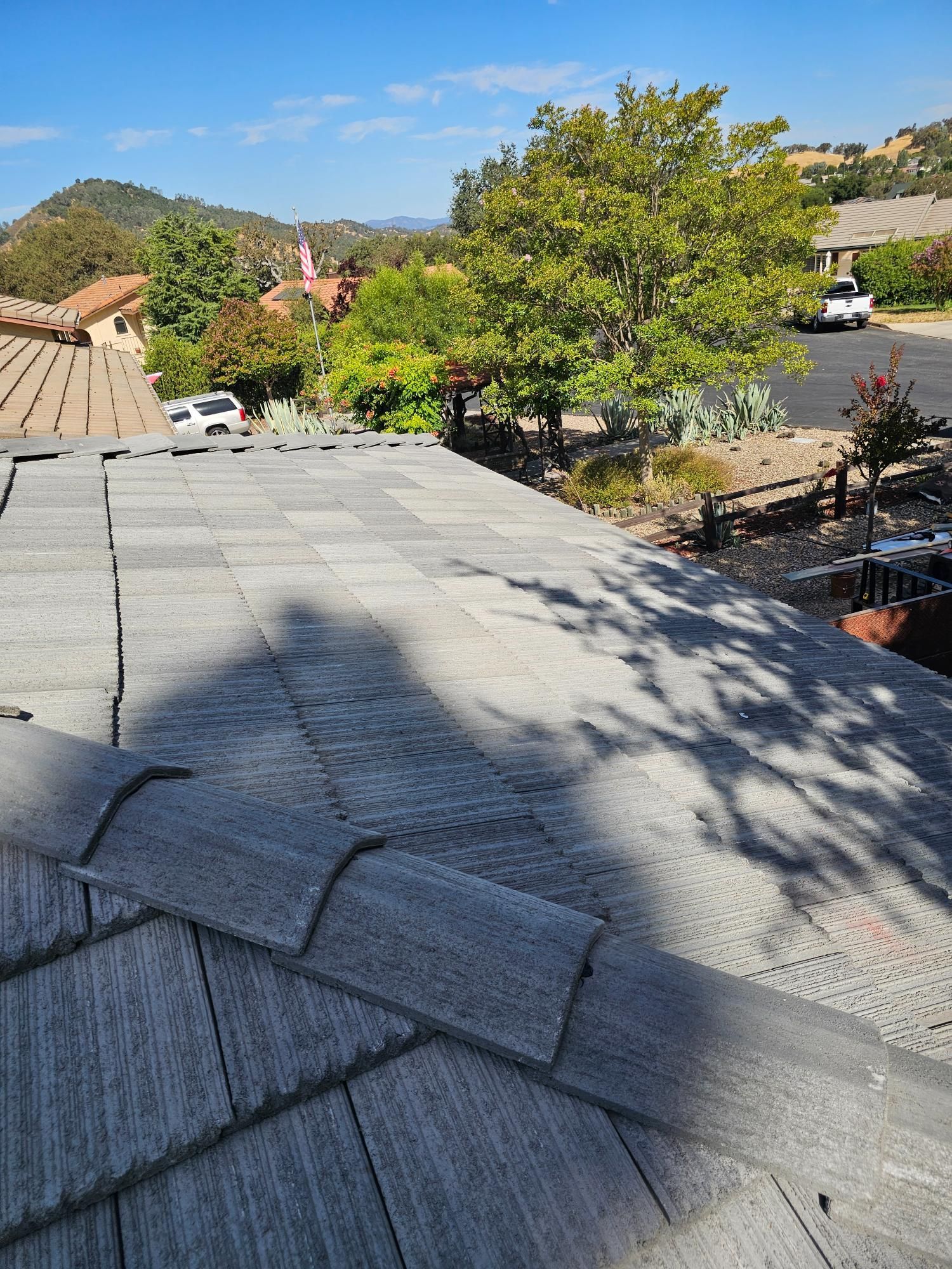 Rooftop shingles with tree shadows, overlooking a sunny residential neighborhood and hills.