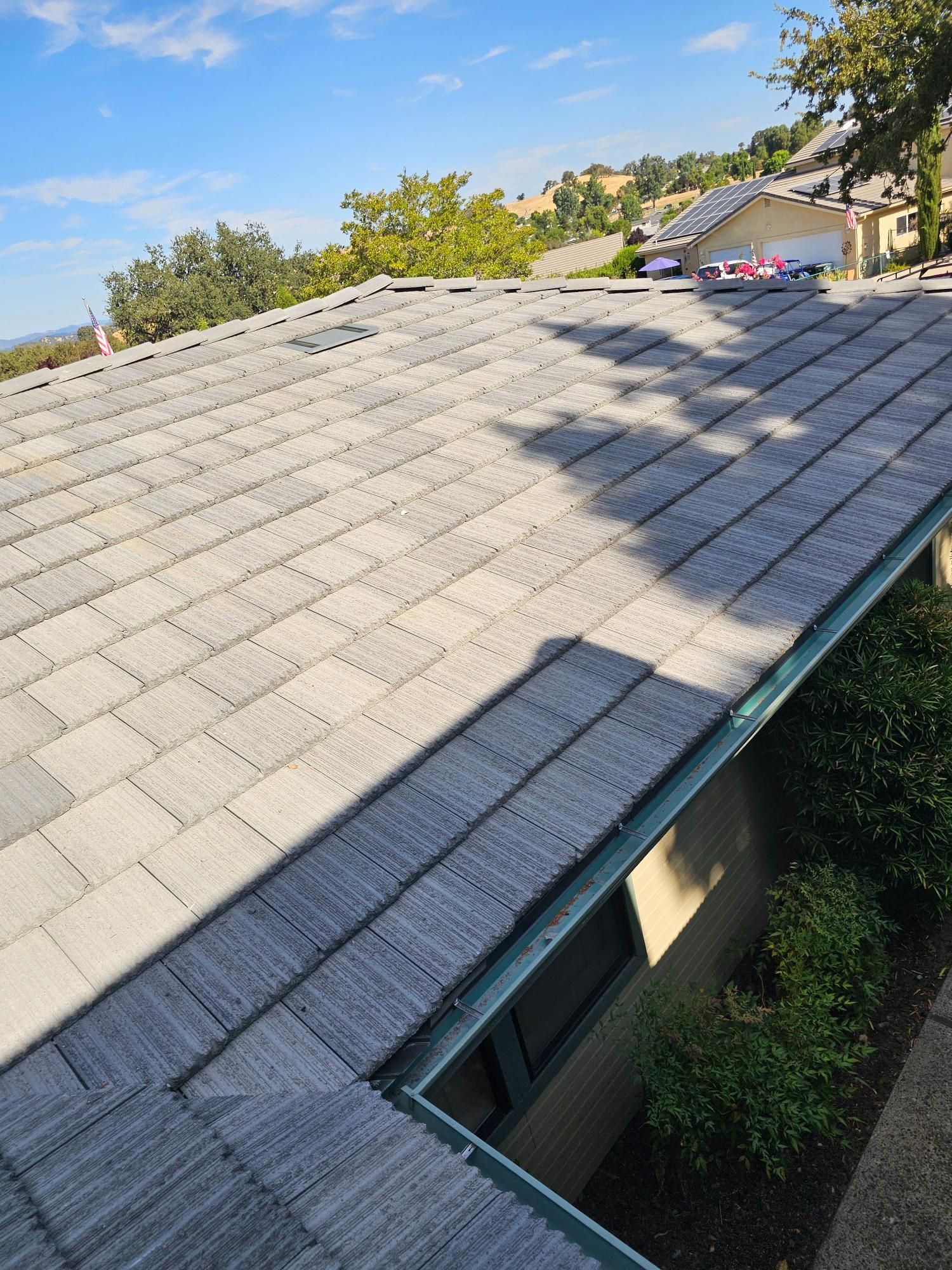 Gray shingle roof with a skylight and tree shadows under a clear blue sky