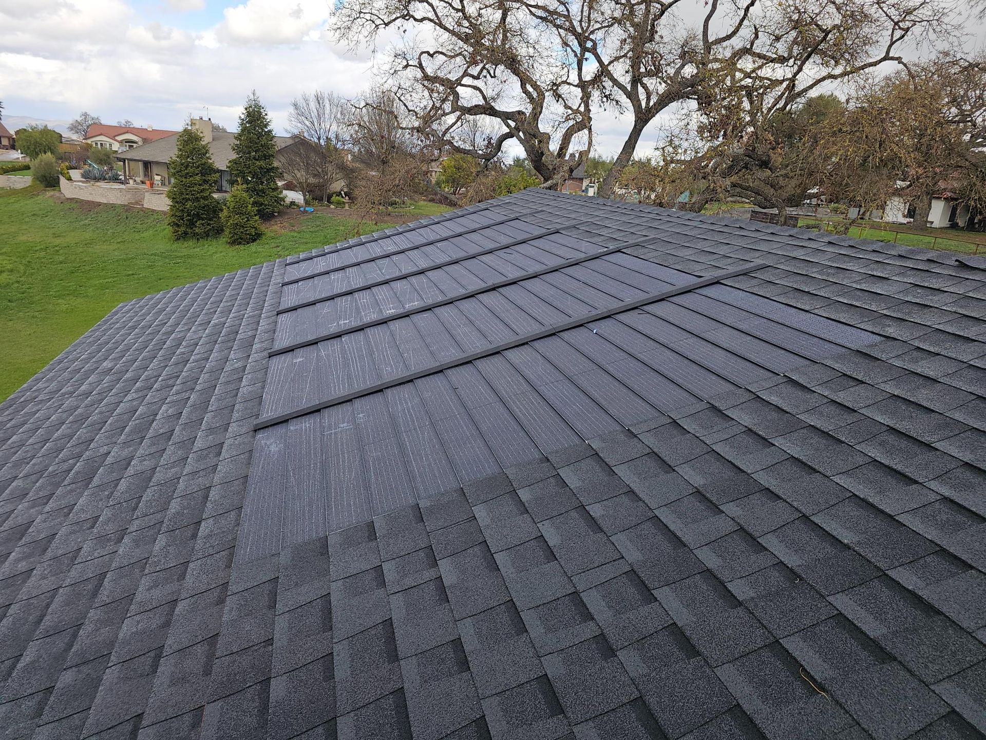 Dark gray shingled roof on a house, viewed from above with trees and grass in the background.