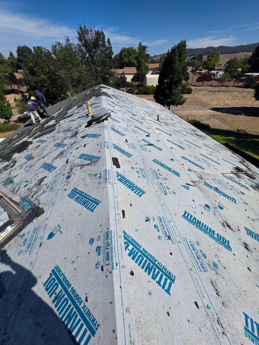 Roof under construction with white underlayment and blue branding, viewed over a tree-lined yard