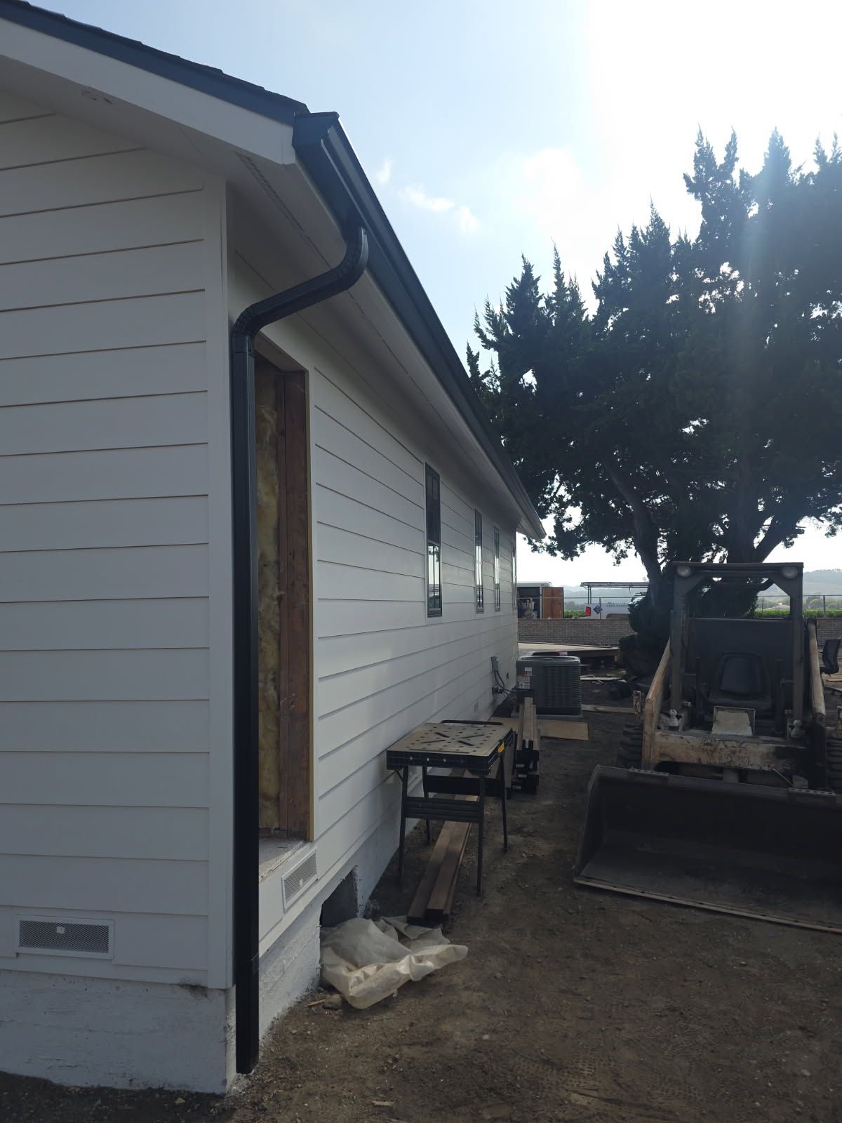 Side of a white house with an open doorway, gravel yard, and a construction tractor near a tree.