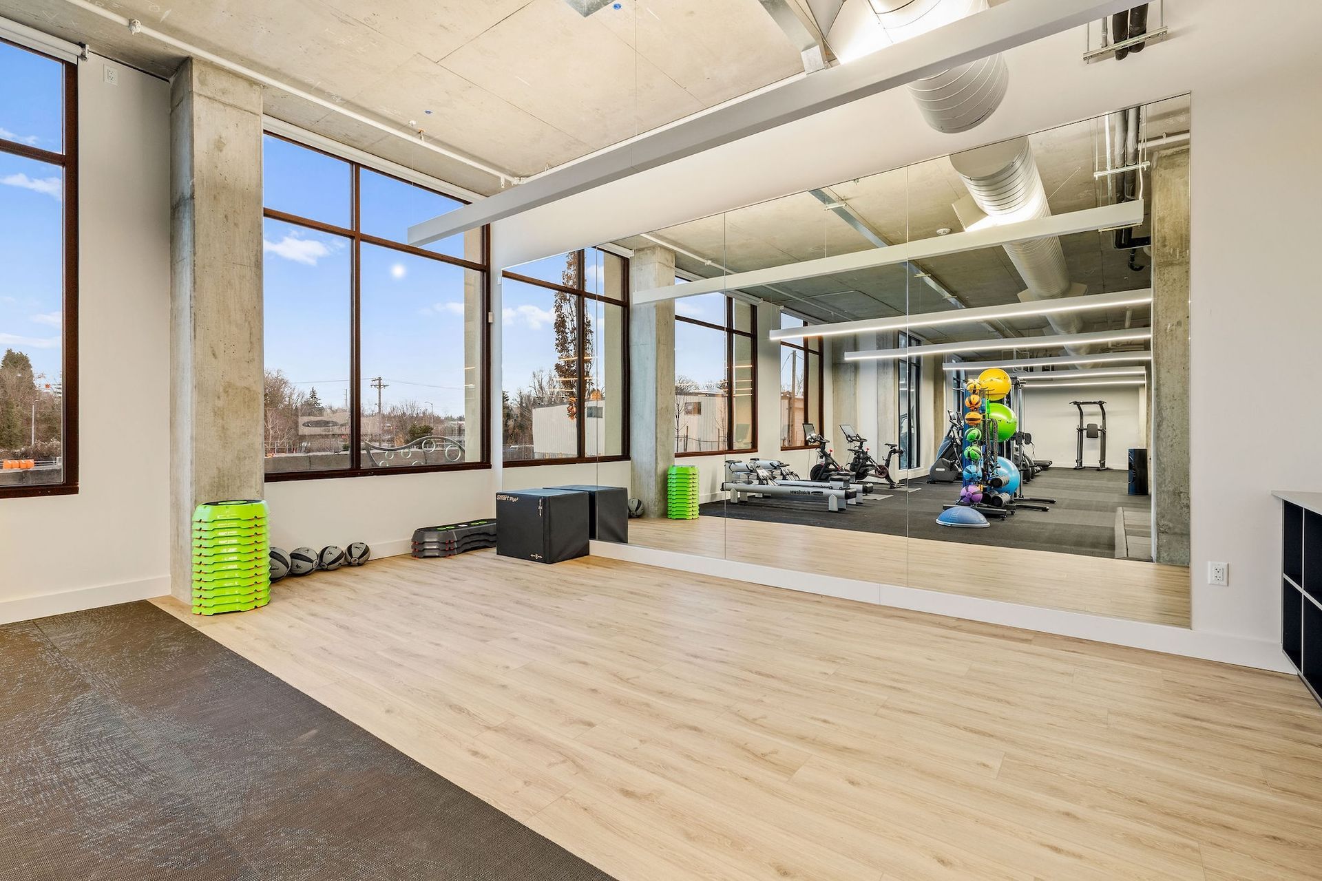 Bright modern gym room with mirrors, windows, wooden floor, and one person exercising on a machine.