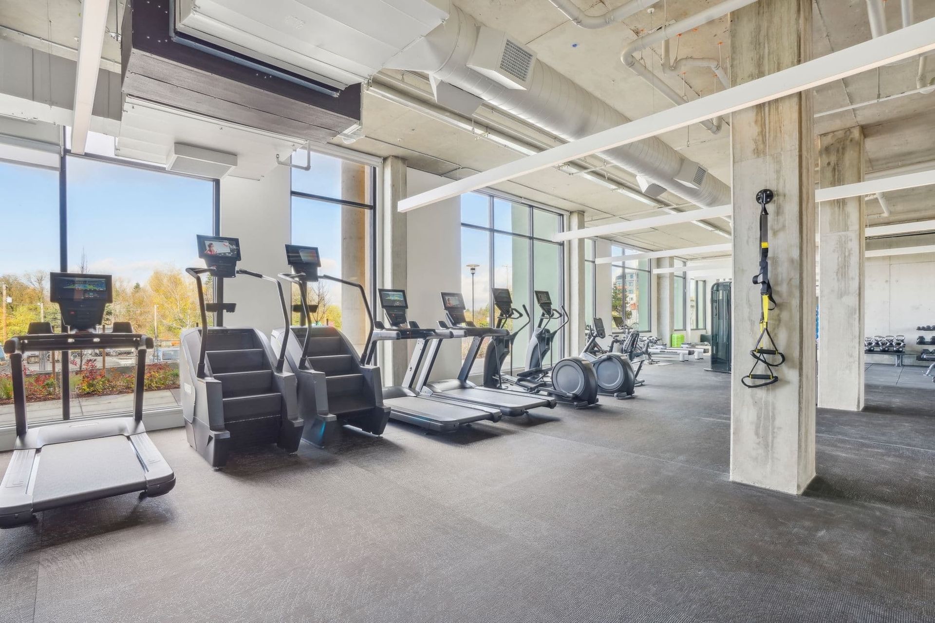 Modern gym with treadmills, ellipticals, rowing machines, and large windows in a bright industrial space