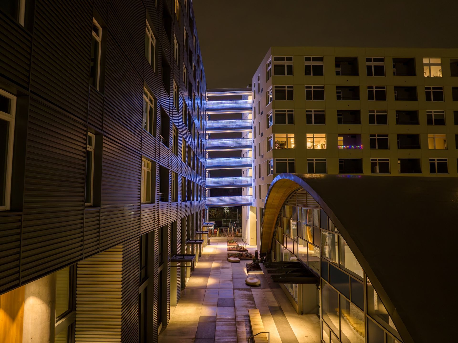 Nighttime urban walkway between buildings, lit by glowing windows and blue-lit stairs.