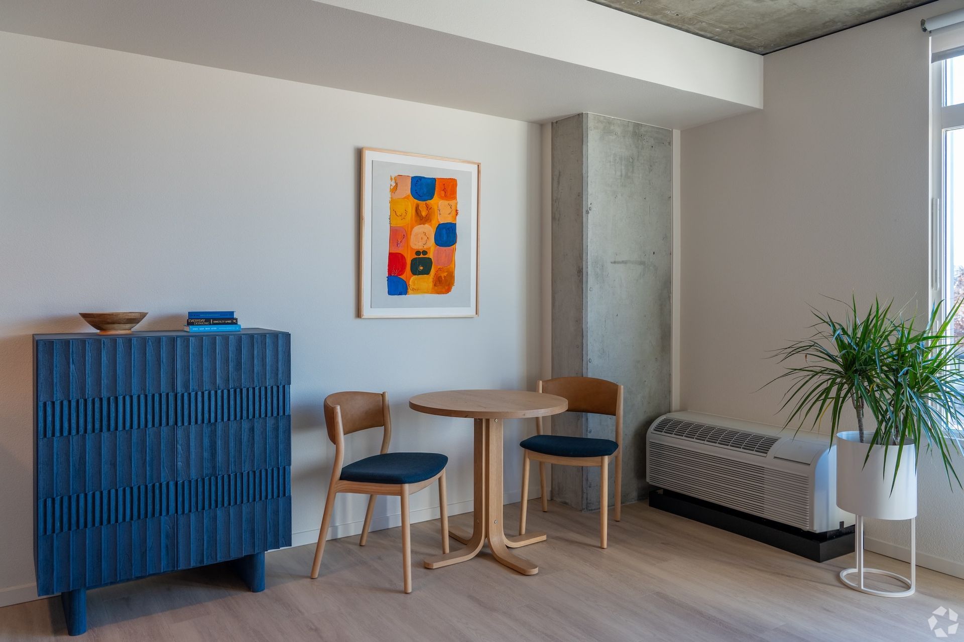 Modern lobby with blue cabinet, small table and chairs, framed art, radiator, and potted plant.