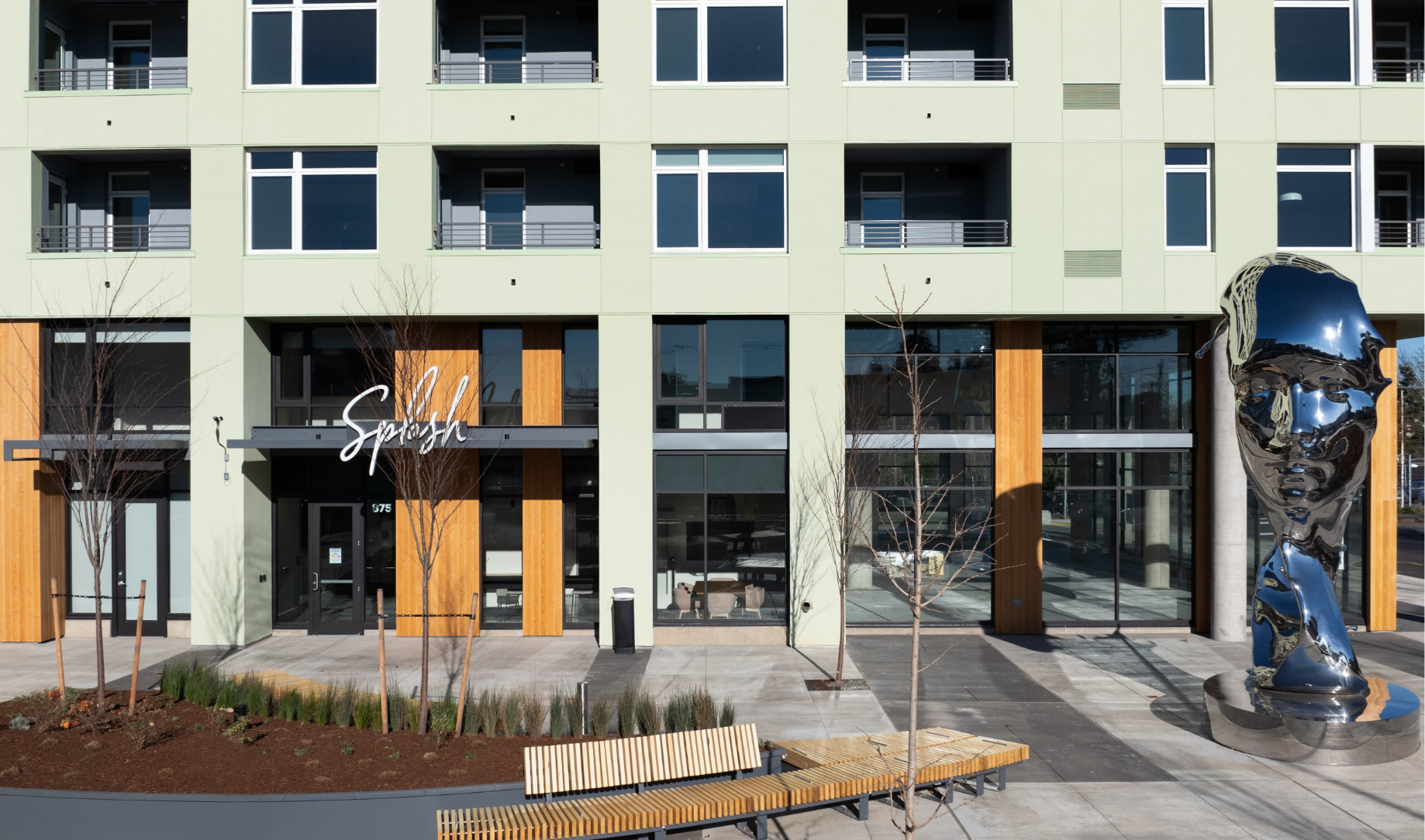 Modern apartment building entrance with glass doors, orange accents, and a blue abstract sculpture in front.
