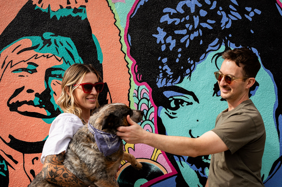Two people smiling and petting a dog in front of a colorful mural.