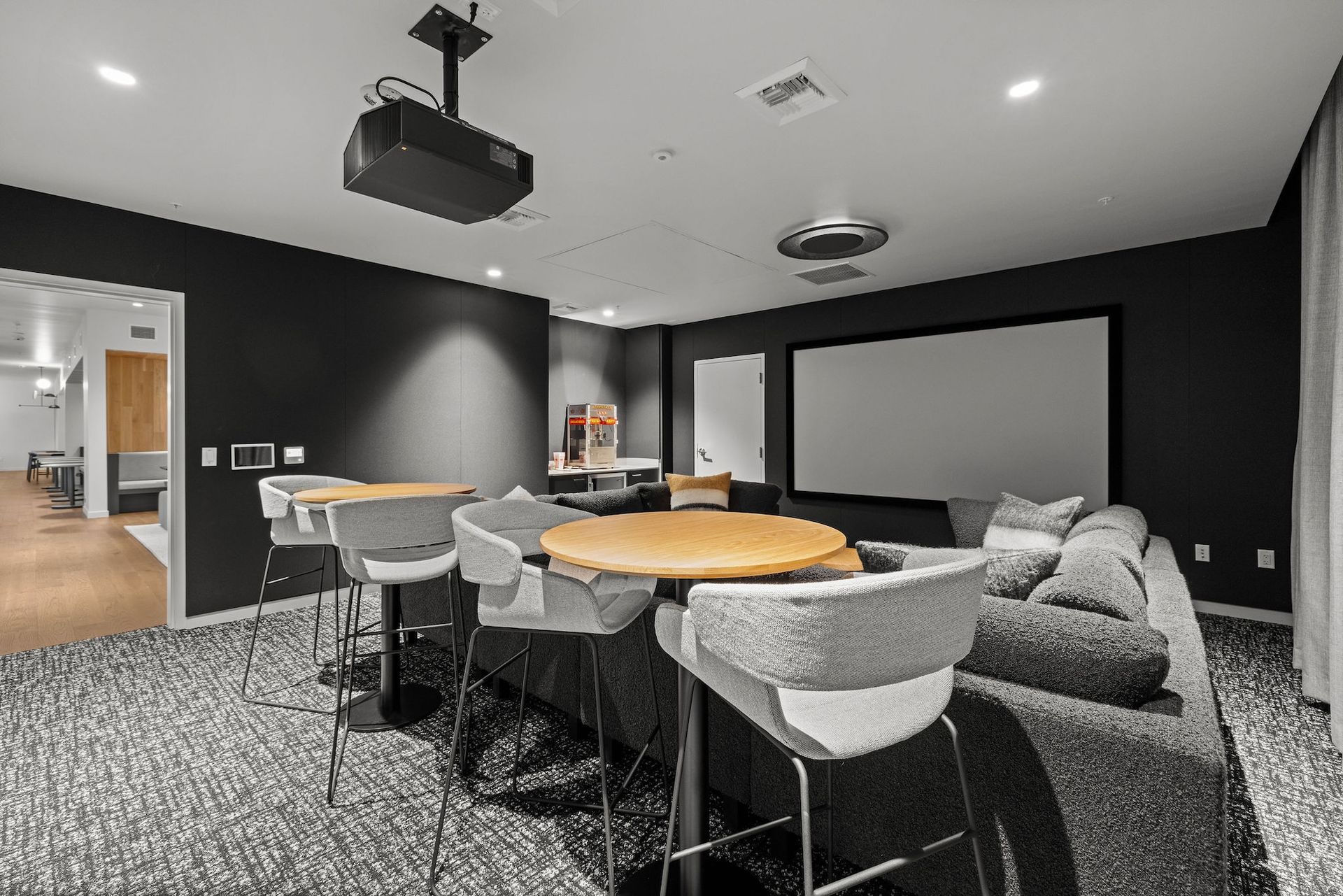 Modern lounge with gray seating, round tables, carpeted floor, and wall-mounted projector screens