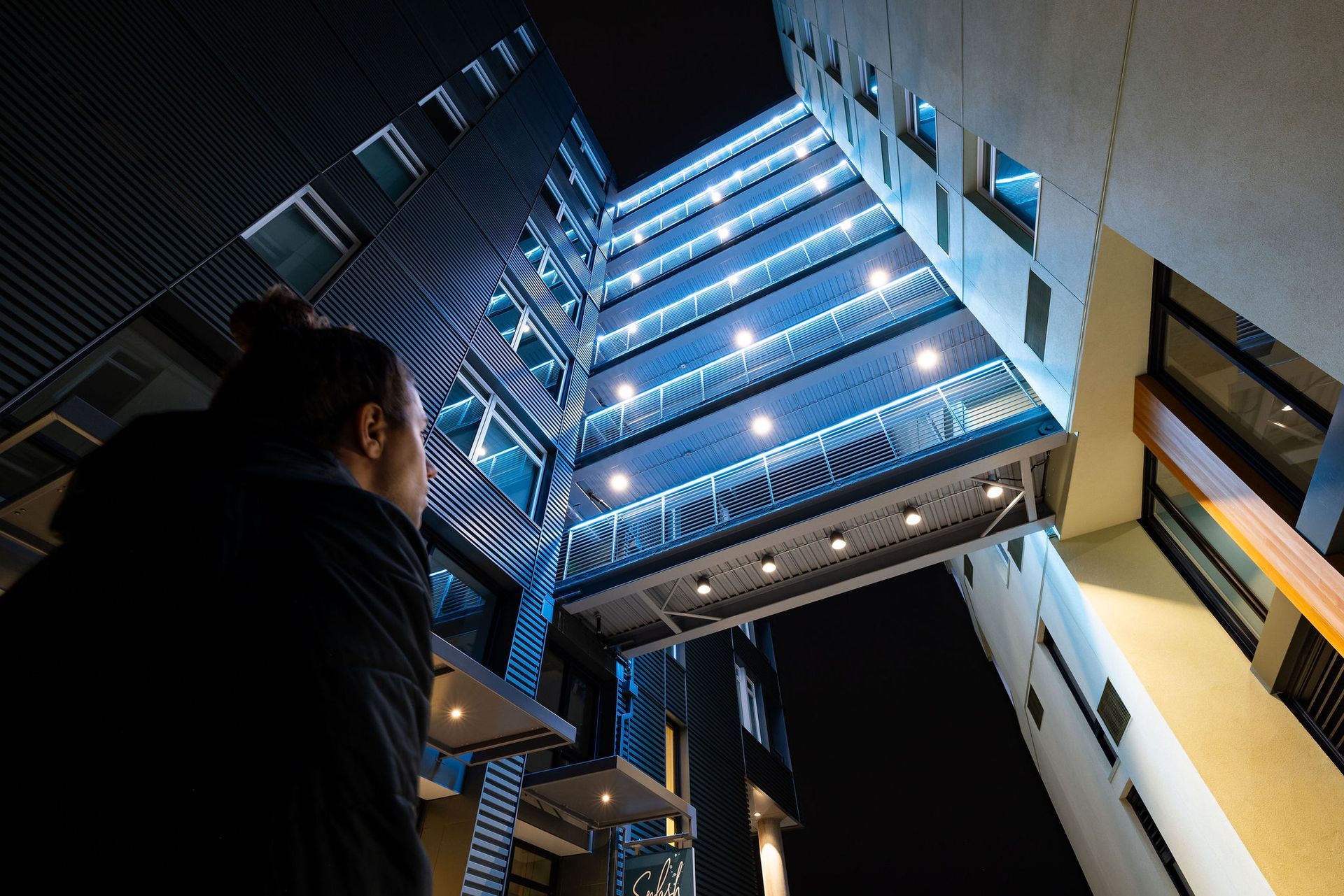 Person looking up at illuminated blue stairwell in a building atrium at night