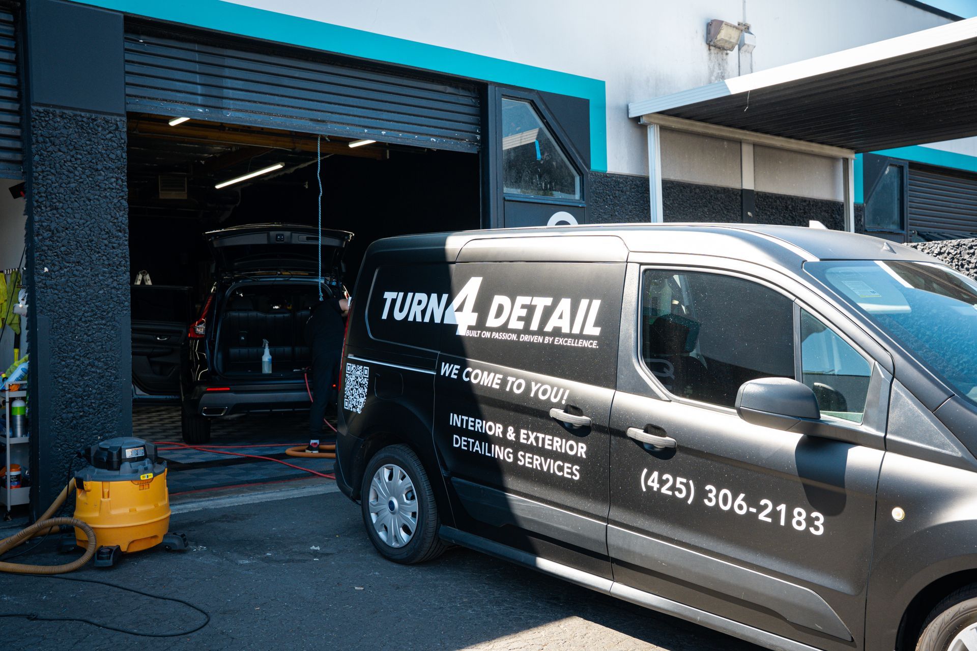 Black detail van parked outside a car wash bay with a visible open garage and cleaning supplies