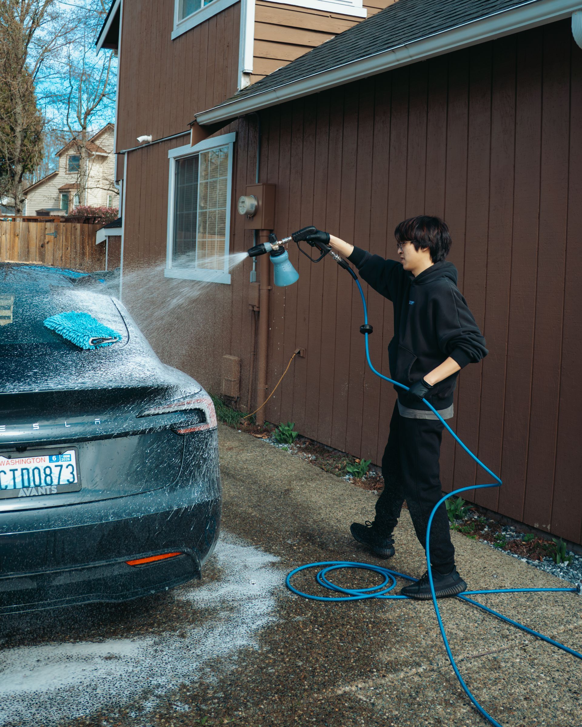 Person pressure-washing a dark car beside a brown house, with water spraying across the driveway.