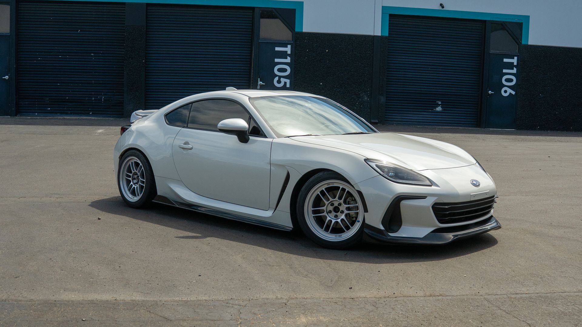 Silver sports car parked on pavement in front of blue shipping containers