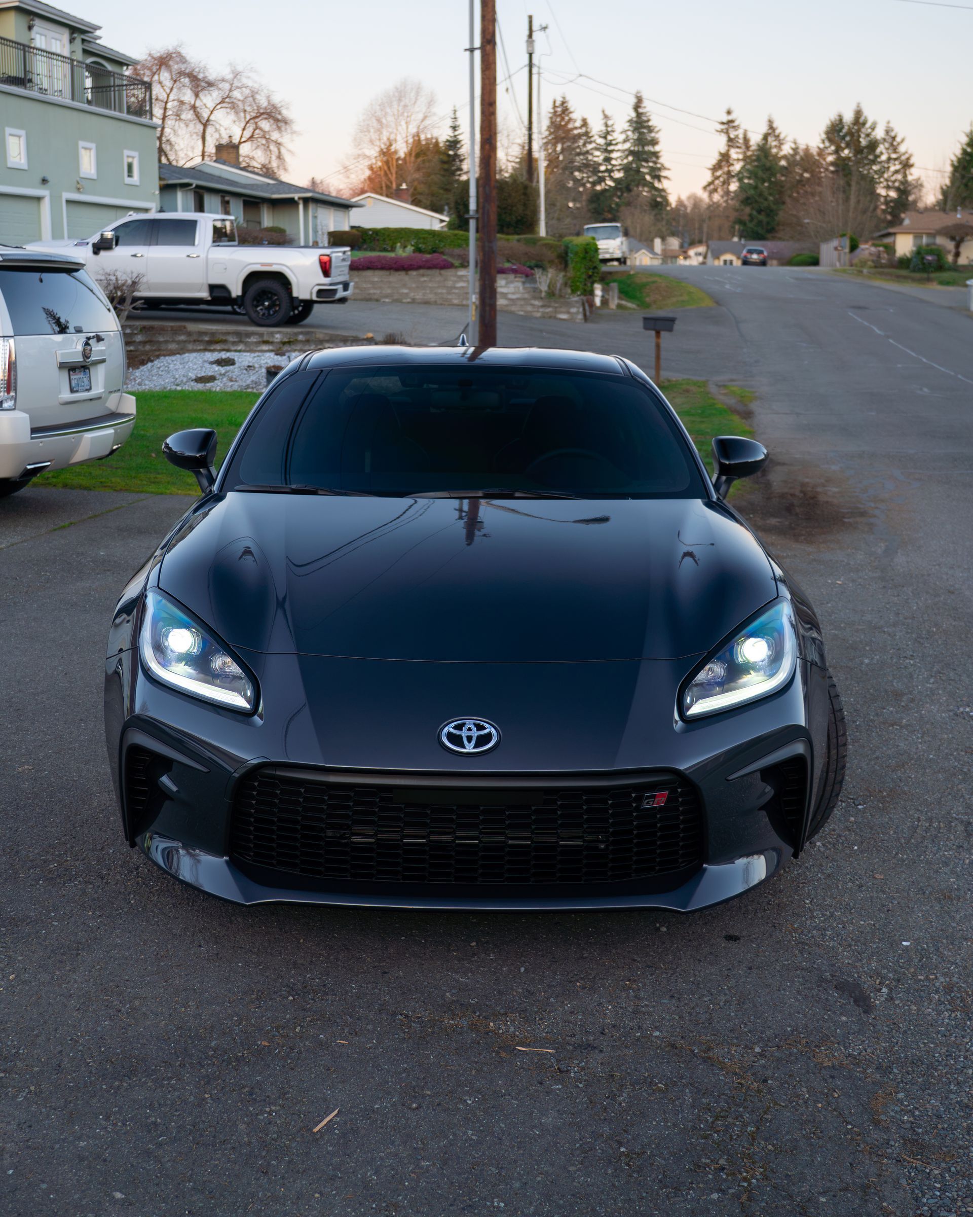 Black sports car parked on a suburban street at dusk, front view with headlights on