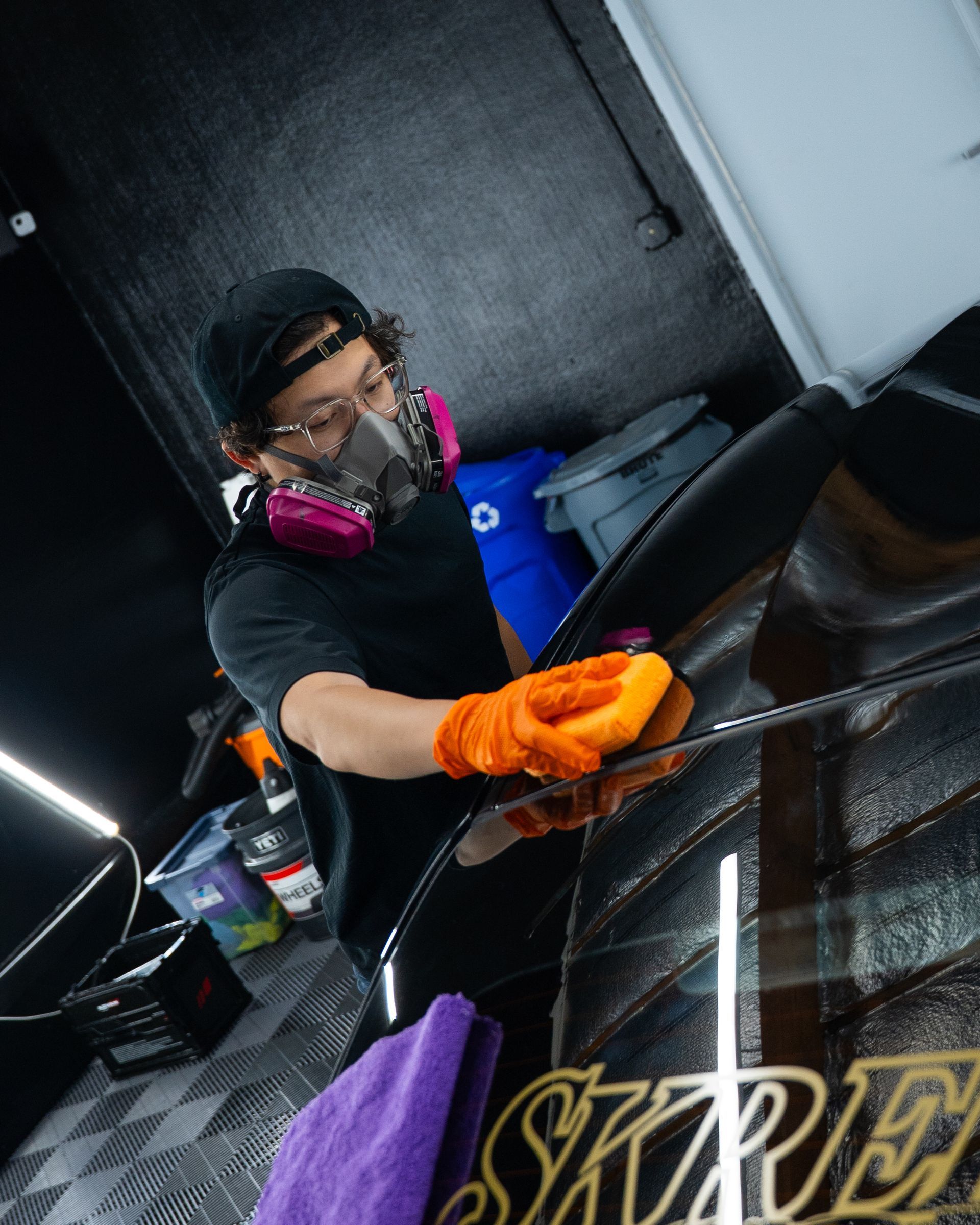 Person in orange gloves polishing a black car in a garage, wearing a respirator and pink hearing protection