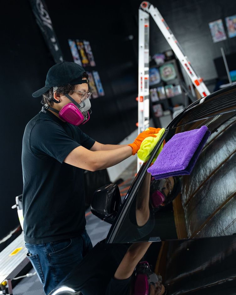 Worker cleaning a black car with a purple sponge and yellow cloth under bright lights