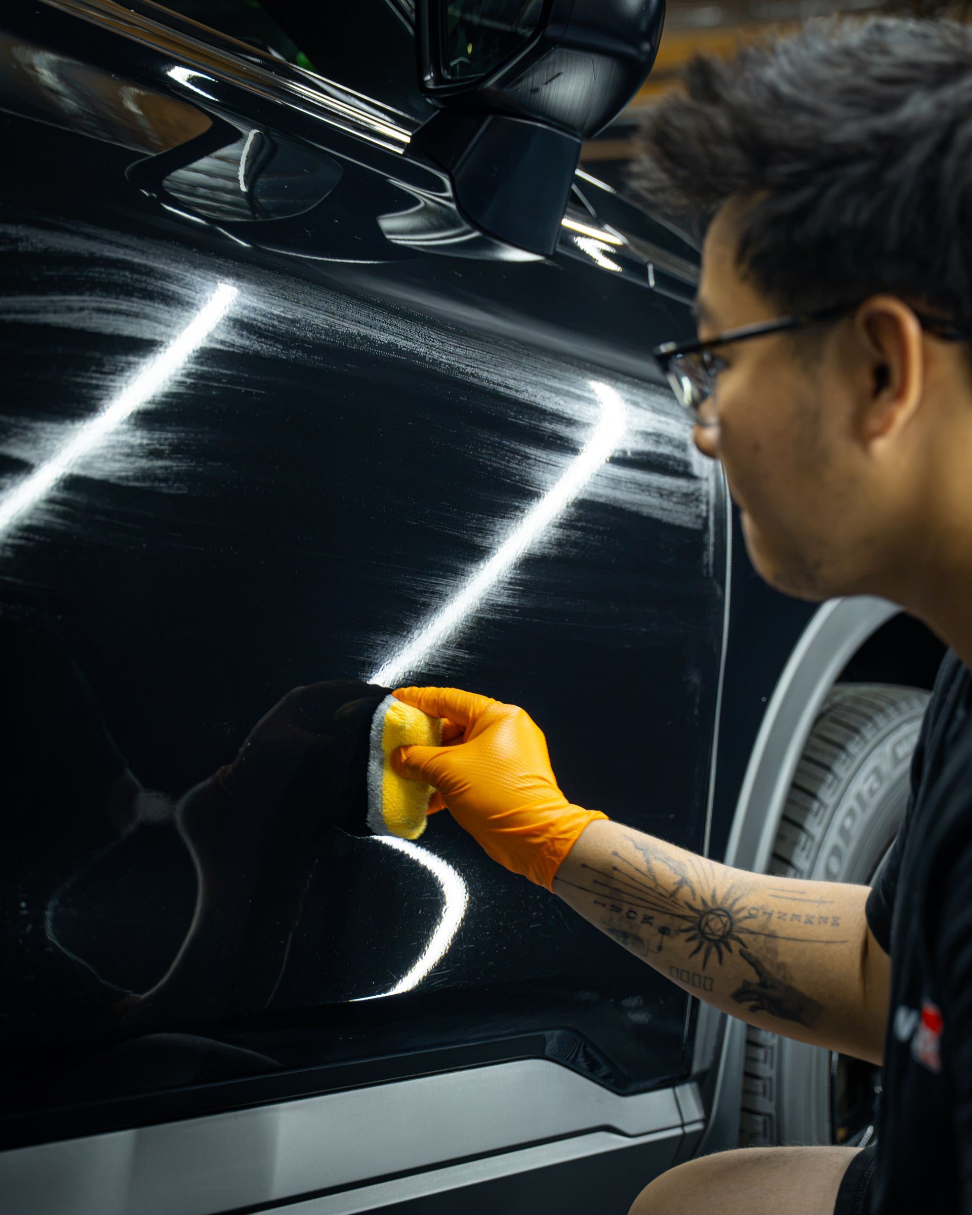 Person wiping a black car door with a yellow cloth in a garage, showing bright reflections on the paint