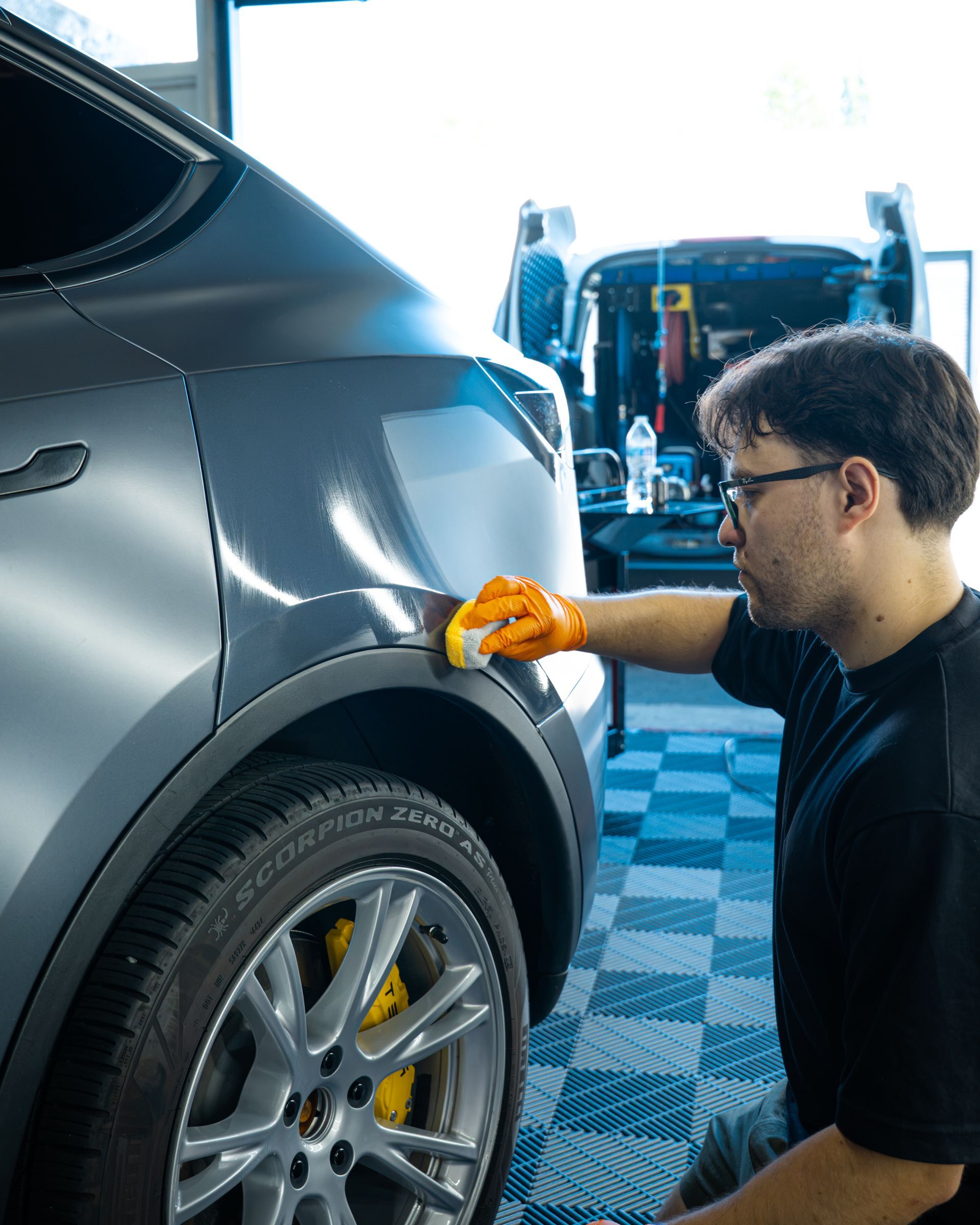 Person polishing a gray car’s rear fender in an auto shop, with a wheel and garage tools visible.