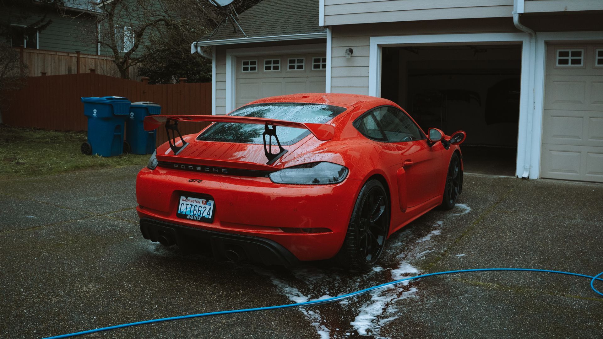 Red sports car parked in a wet driveway by a garage, with water on the ground and a house in the background