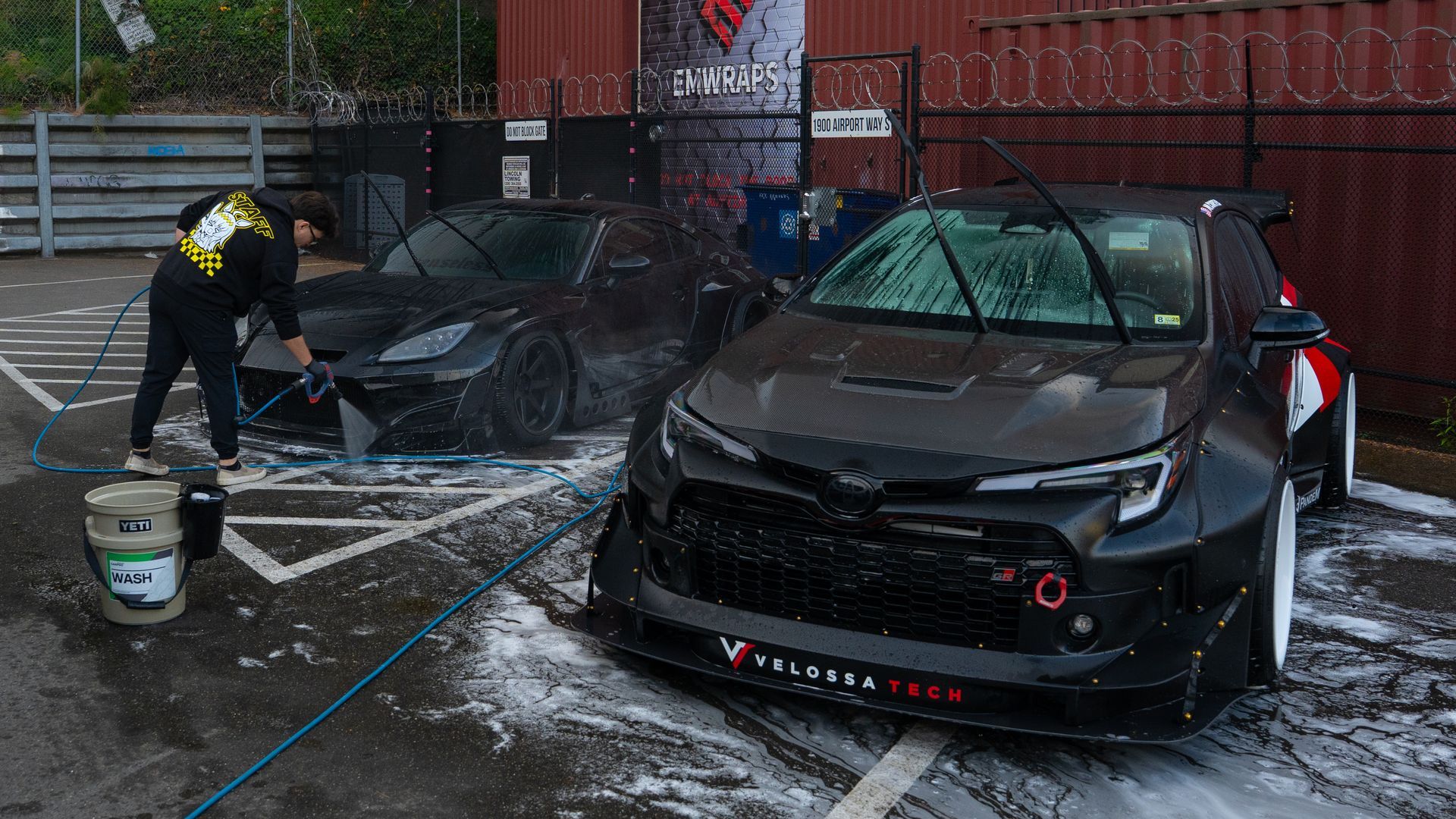 Person washing a black car in a snowy driveway, with another black car nearby.