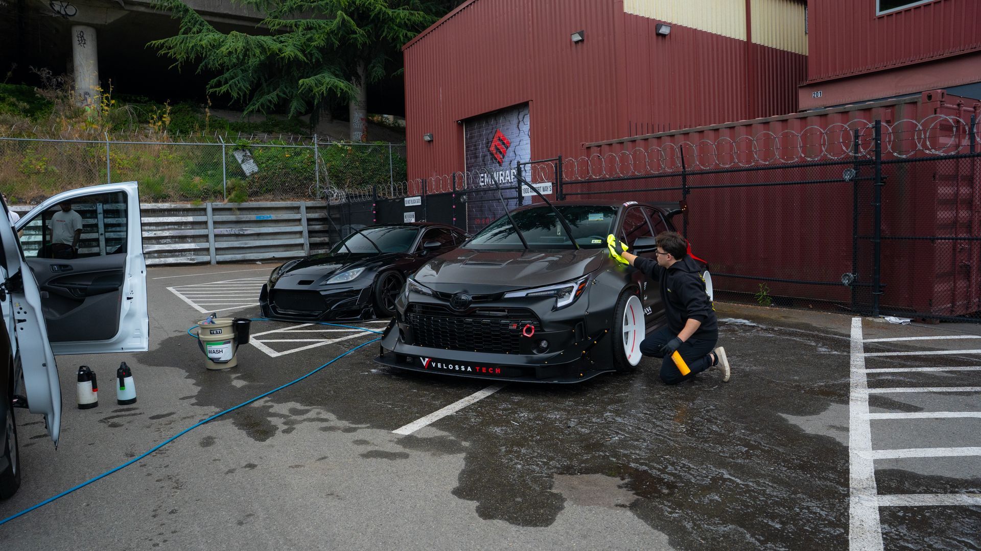 Two black cars in a wet parking lot, with two people standing near one car by a red building.