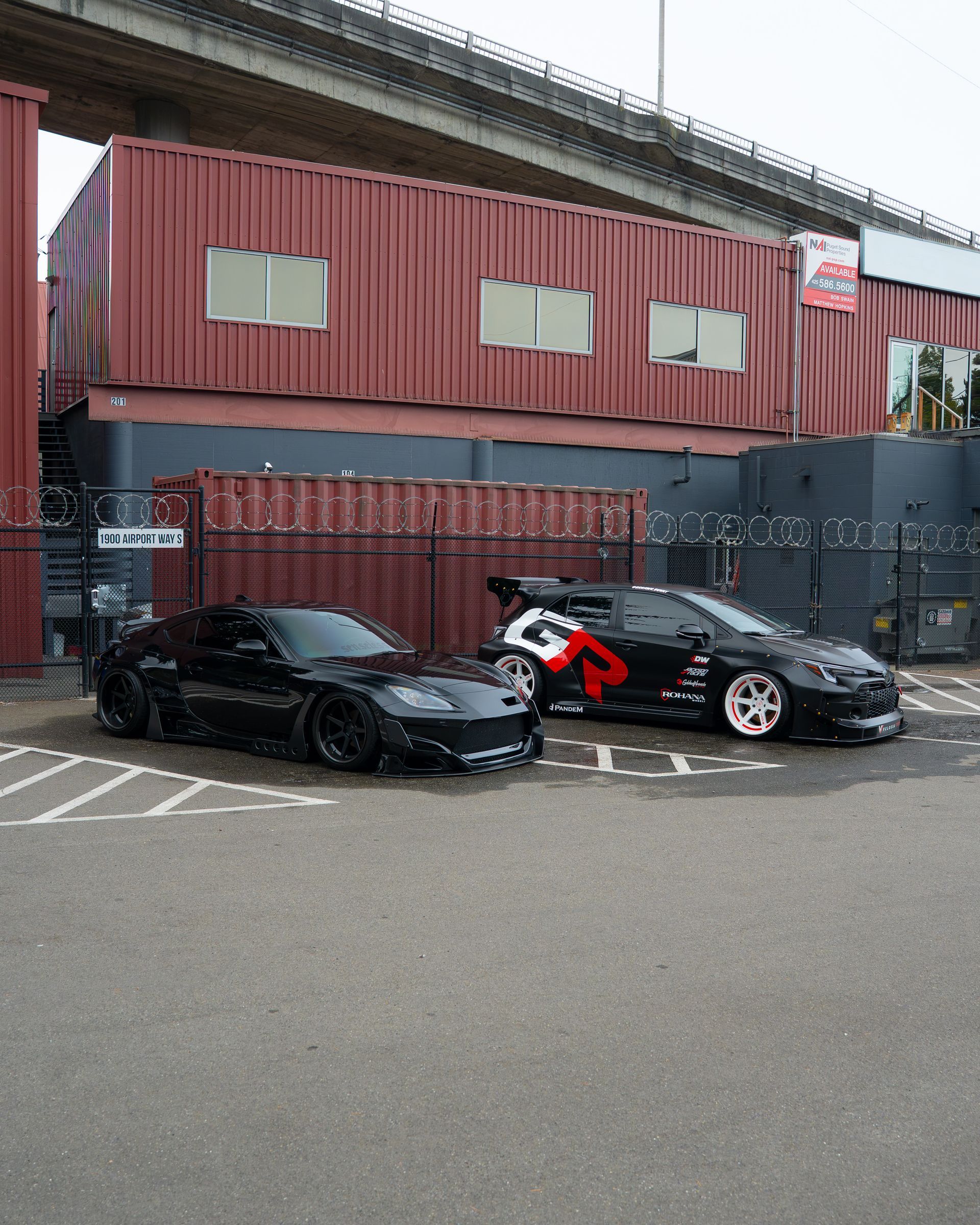 Two black sports cars parked beside a red industrial building with graffiti-covered walls.