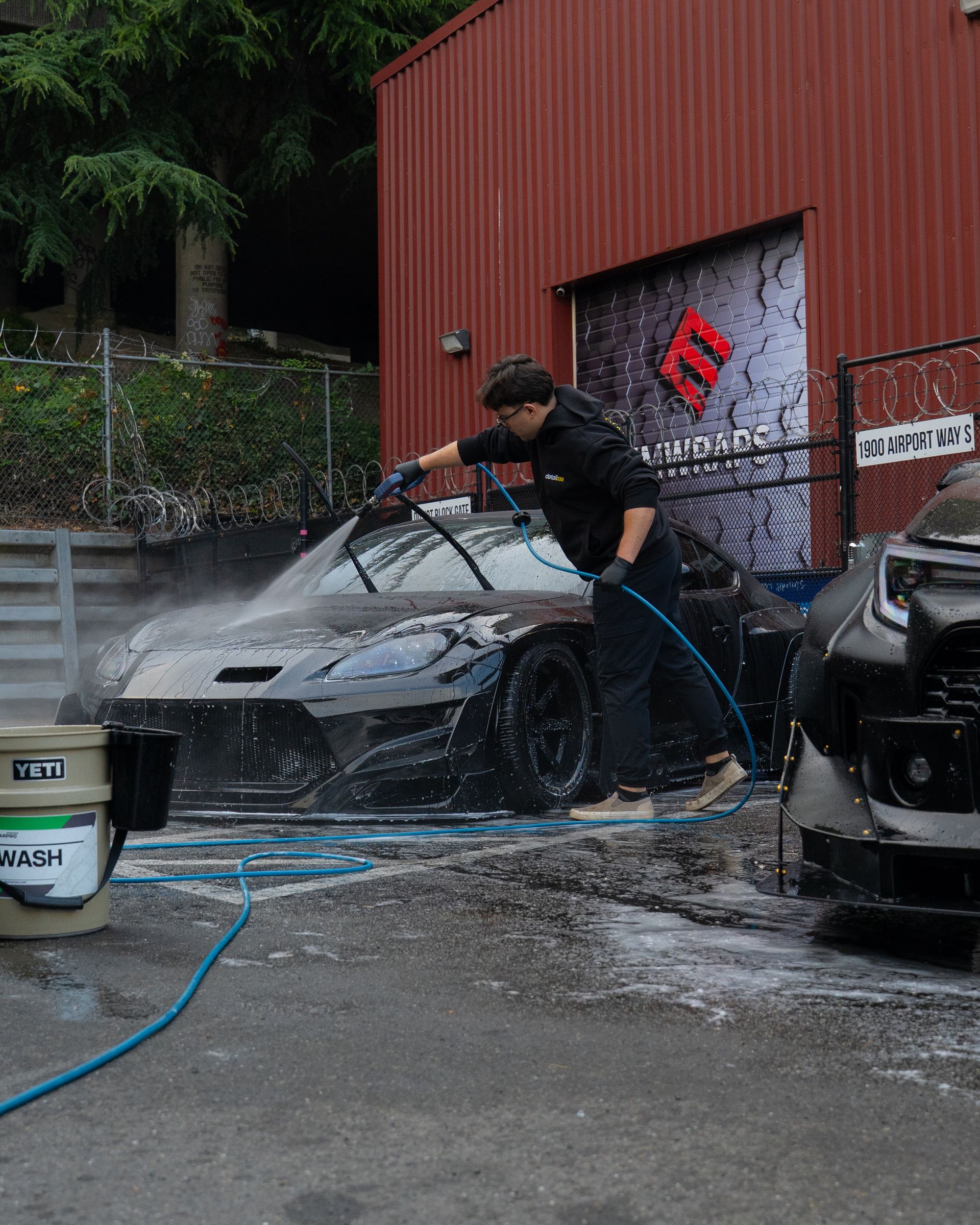 Person pressure-washing a black car in a wet outdoor wash area beside a red building.
