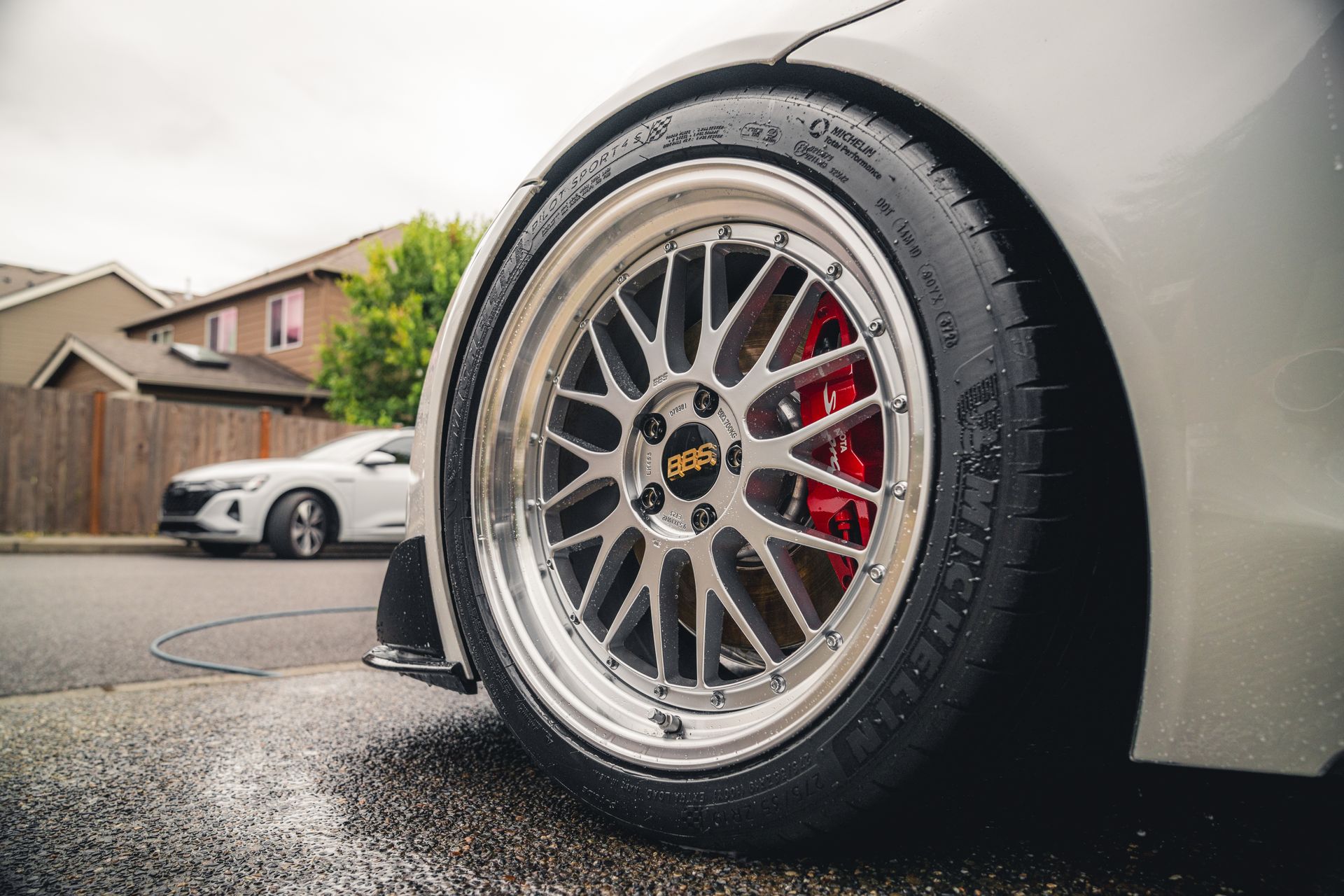 Close-up of a silver sports car wheel with red brake calipers on a wet driveway, another car in the background