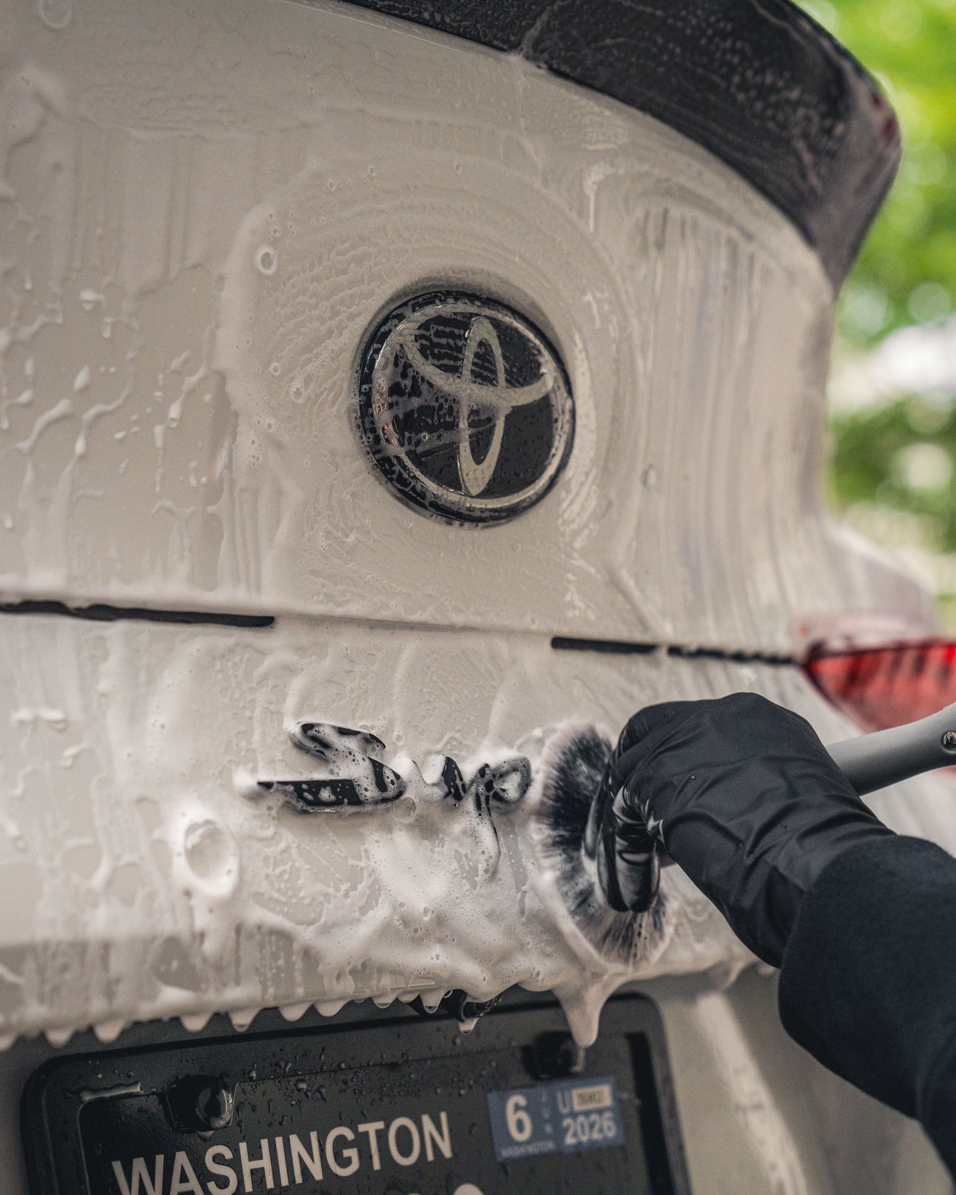 Hand washing a white Toyota SUV with soap foam and a black license plate in a car wash