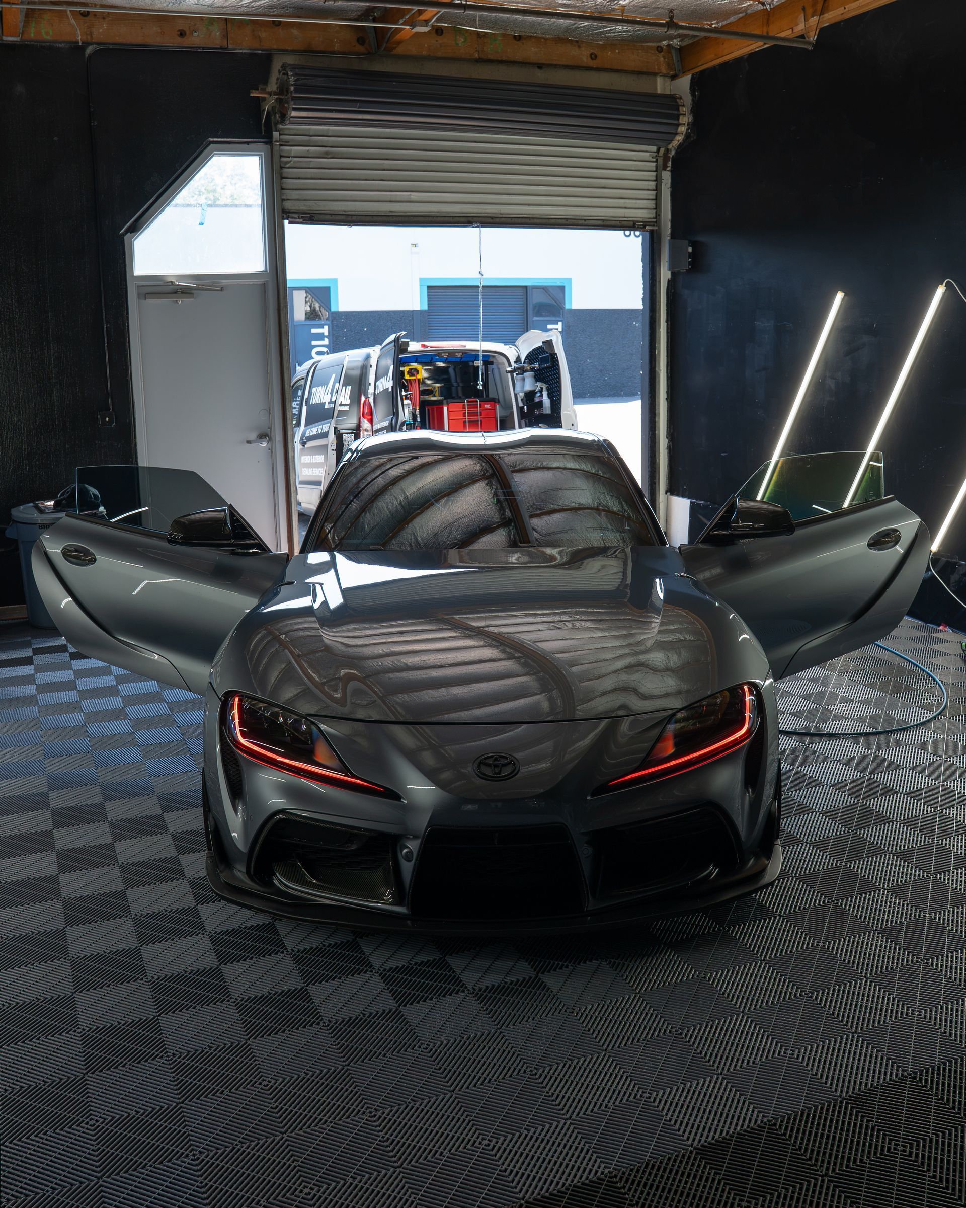 Gray sports car with doors open in a garage, viewed from the rear.