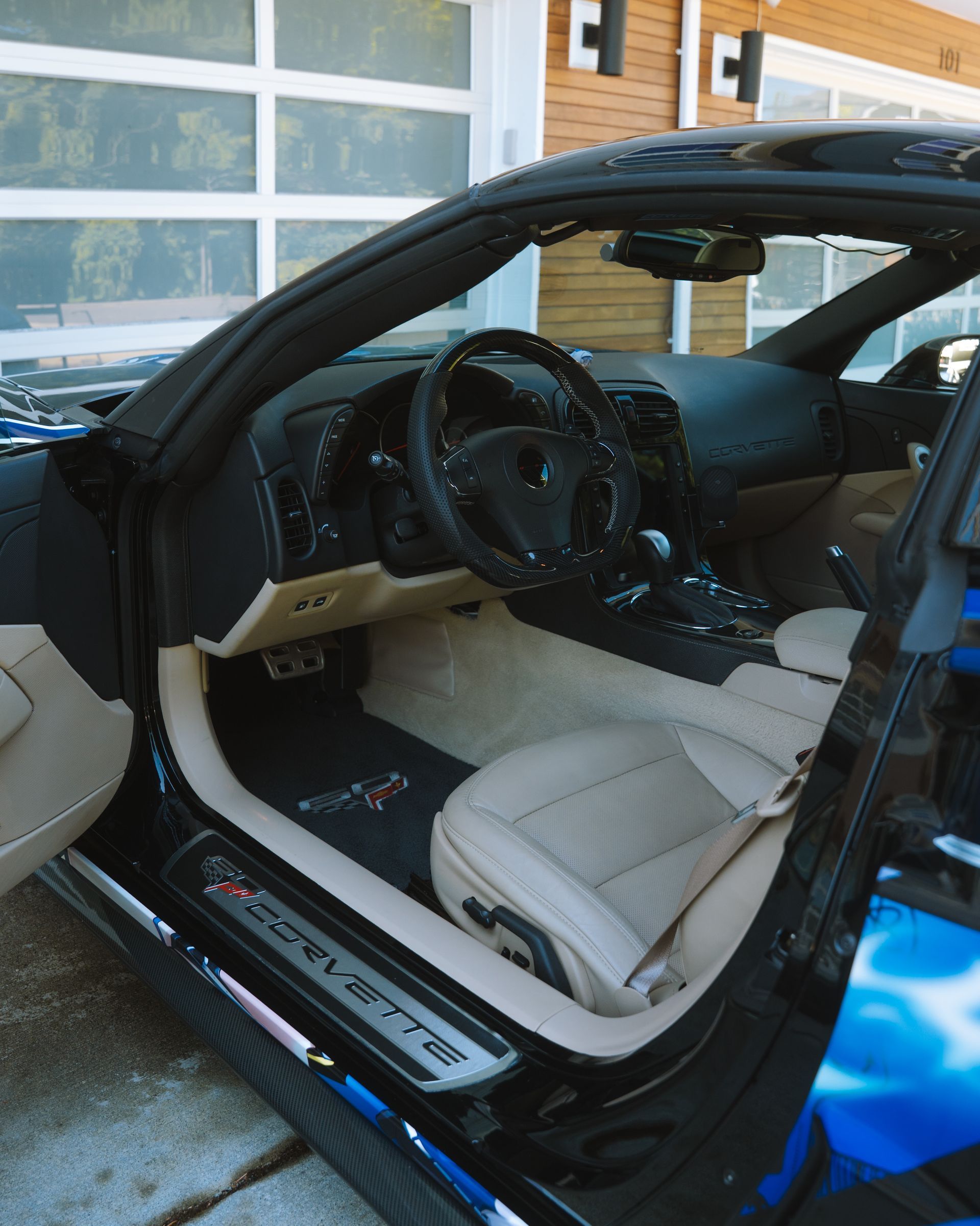 Open car door showing a beige-and-black interior with steering wheel and seat against a garage backdrop