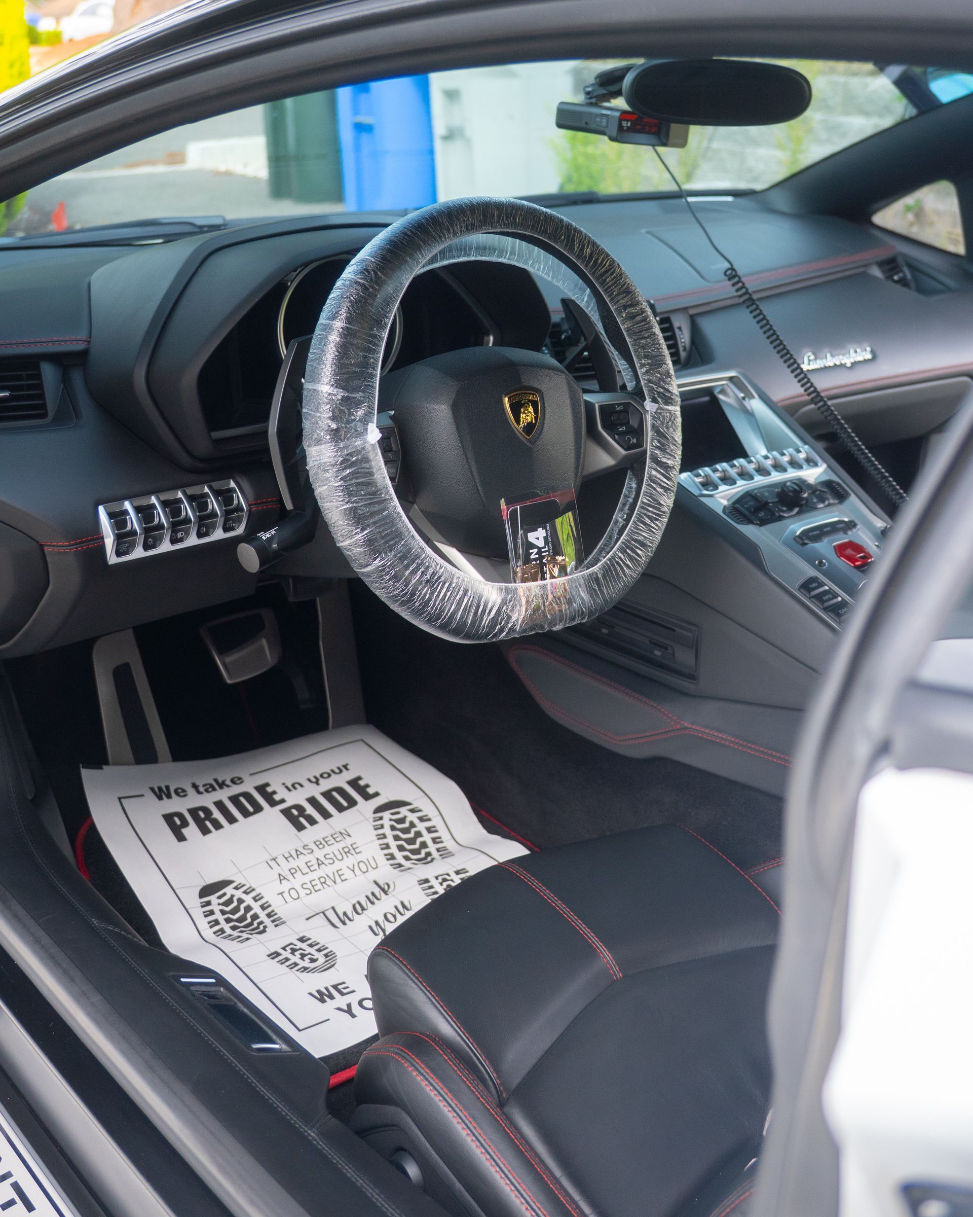 Lamborghini interior with steering wheel wrapped in plastic and a Pride Ride floor mat visible.