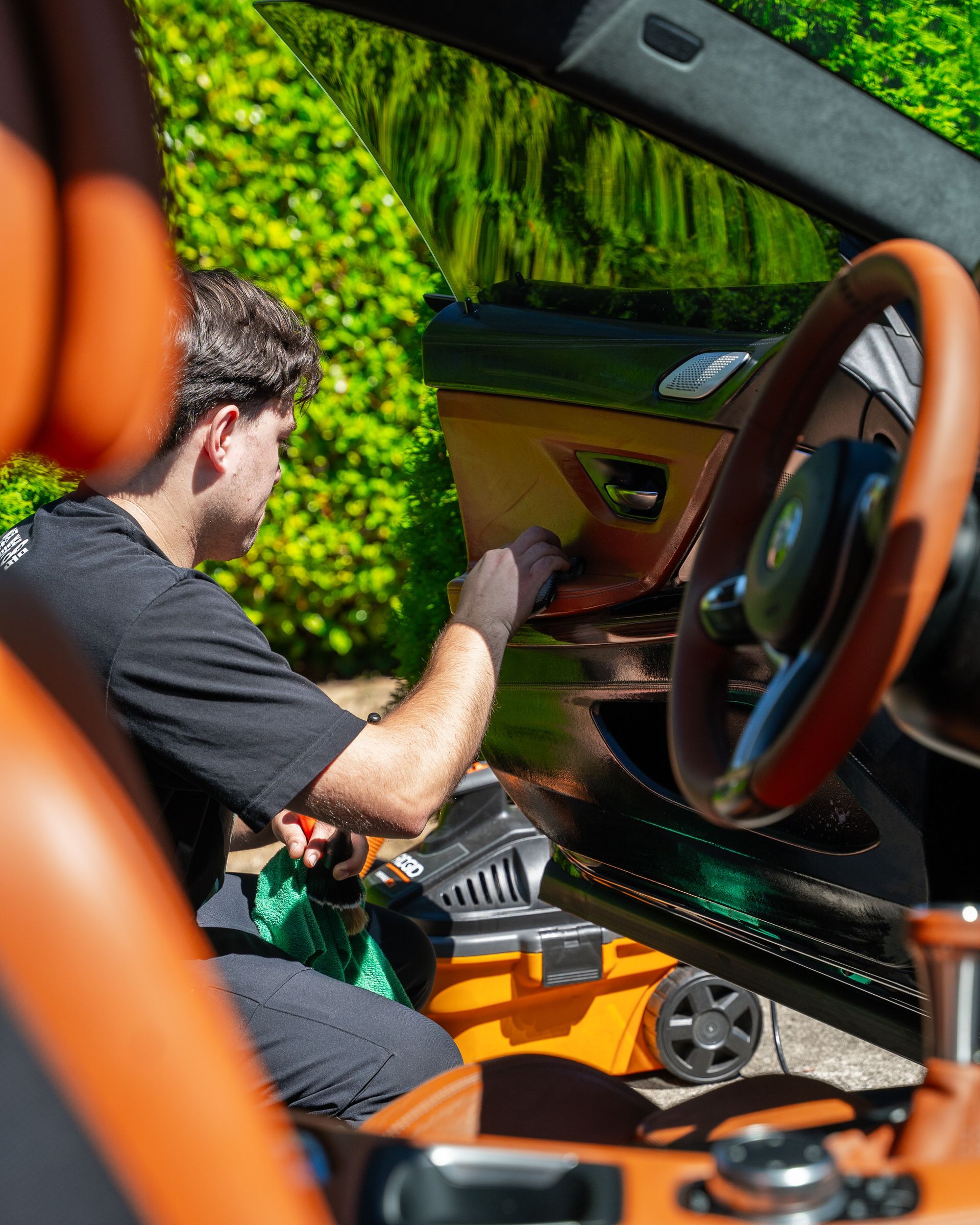 Person cleaning the interior of an orange sports car with a green hedge reflected on the windshield