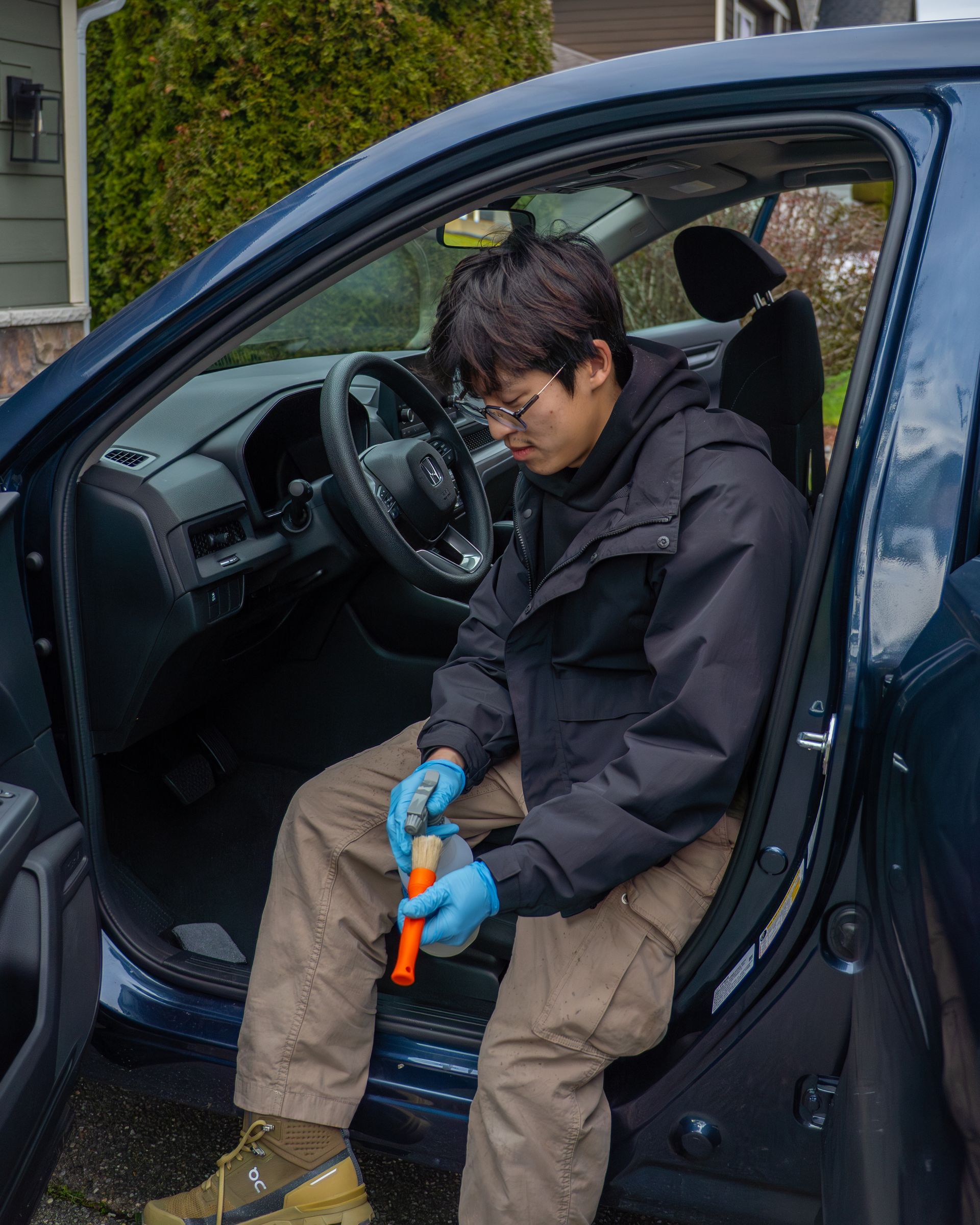Person seated in a car doorway holding a blue-and-orange tool, wearing gloves and work clothes.