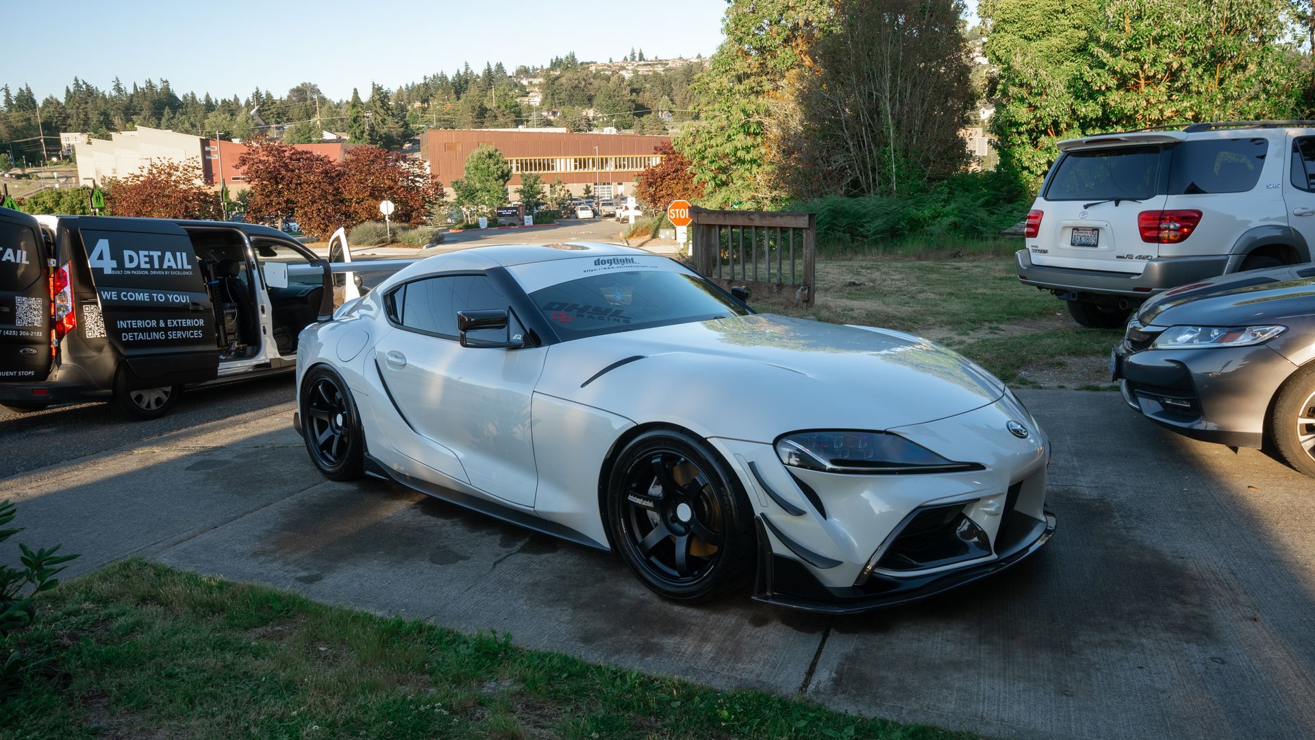 White sports car parked outdoors on a driveway, with other vehicles nearby and trees in the background