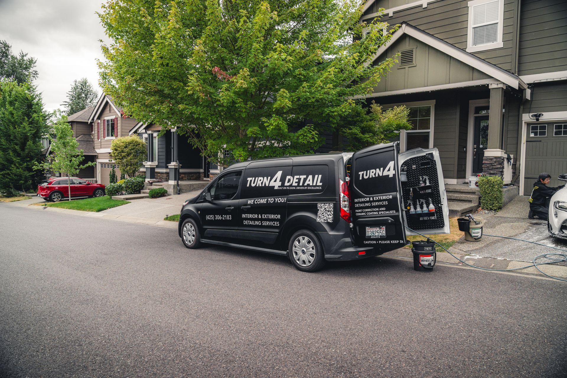 Black delivery van parked on a residential street in front of a house, with open rear doors and signboard.