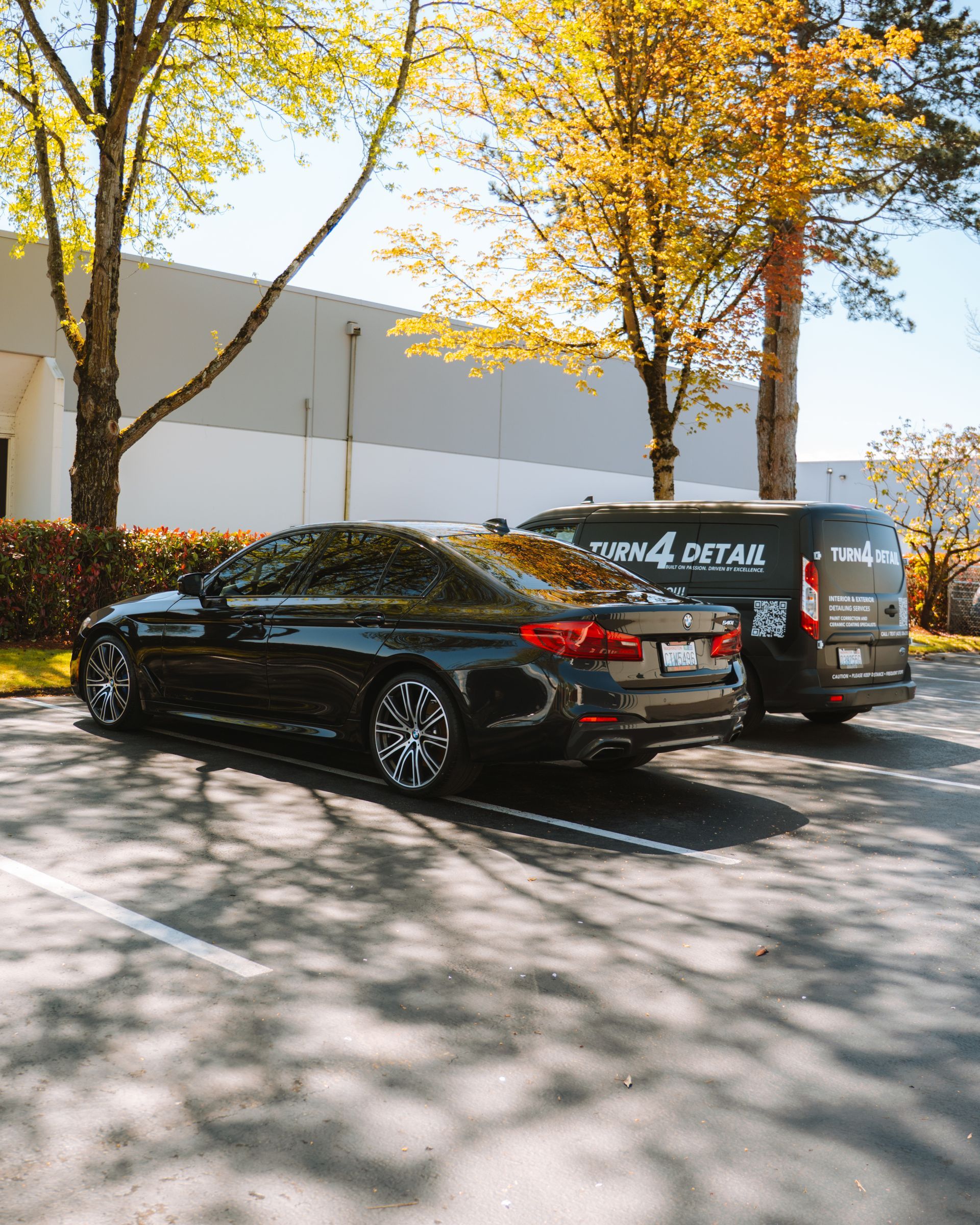 Black sedan and gray van parked beside a white building under autumn trees in sunlight