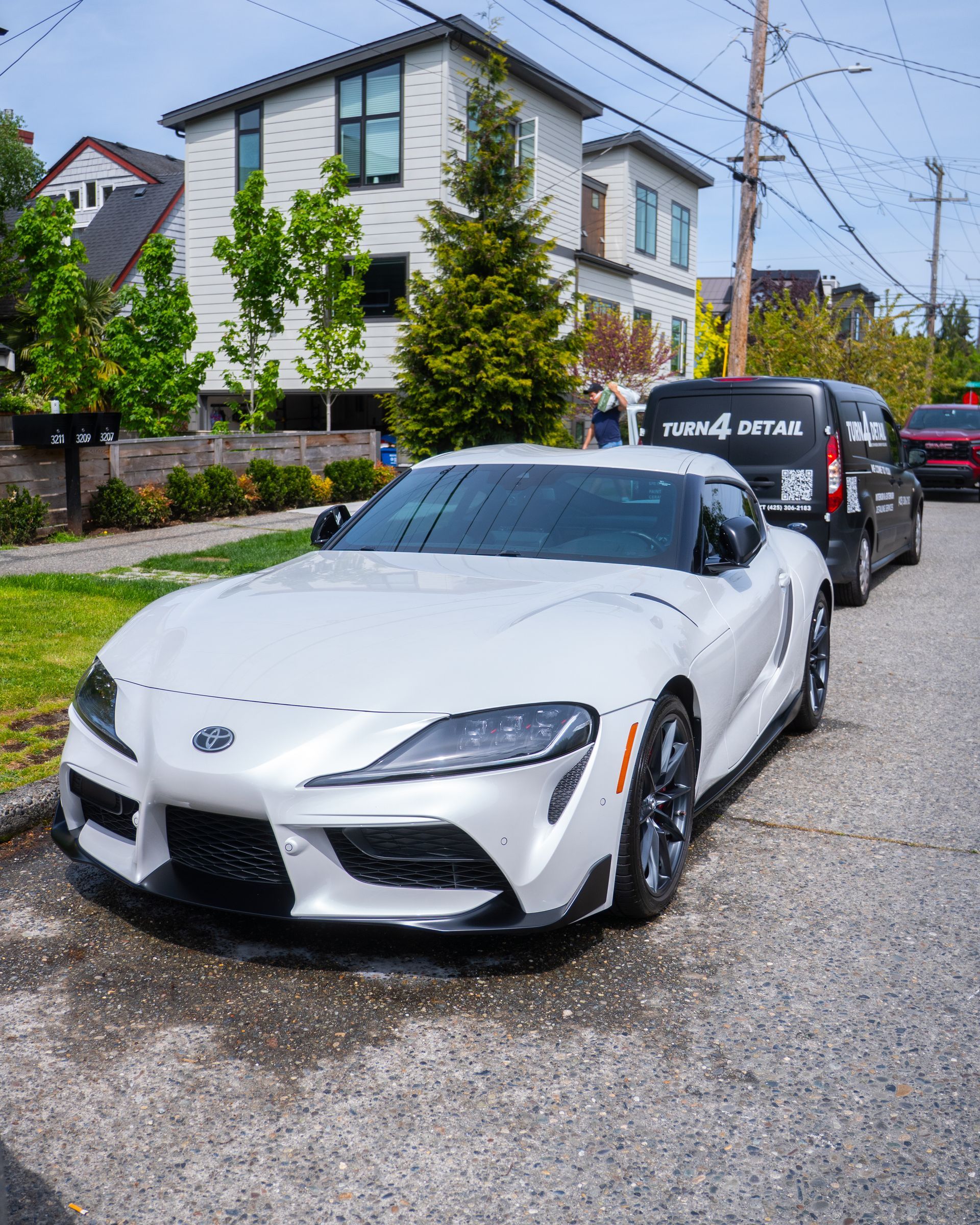 White sports car parked on a gravel driveway beside a black van in front of a house.