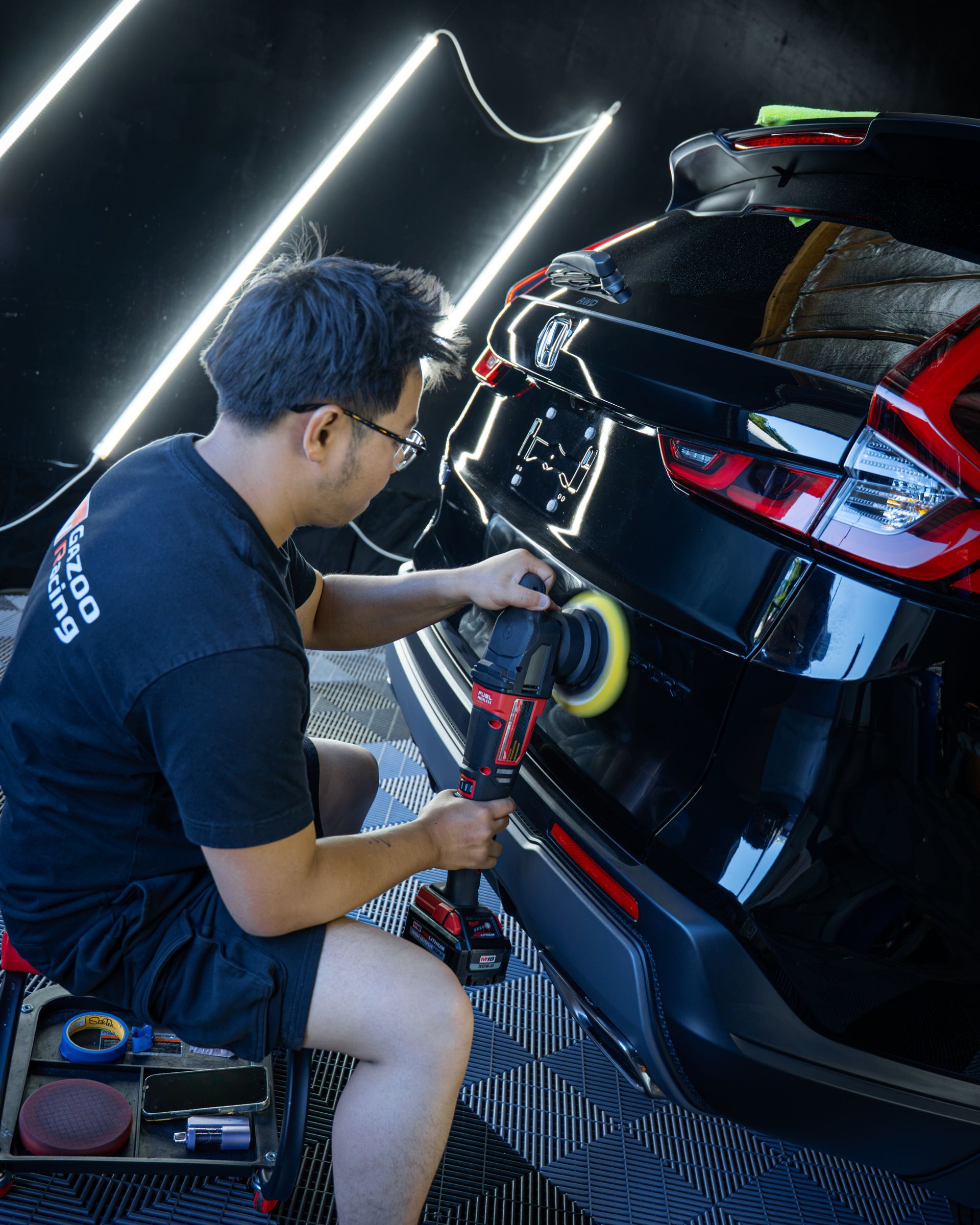 Detailer polishing the rear of a black car with a buffer in a garage
