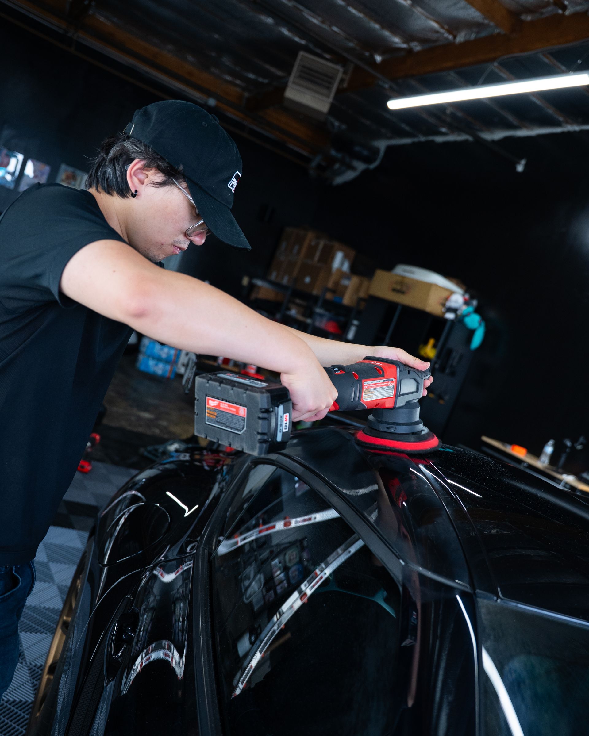 Person polishing a black car hood with a red-and-black power buffer in a garage.