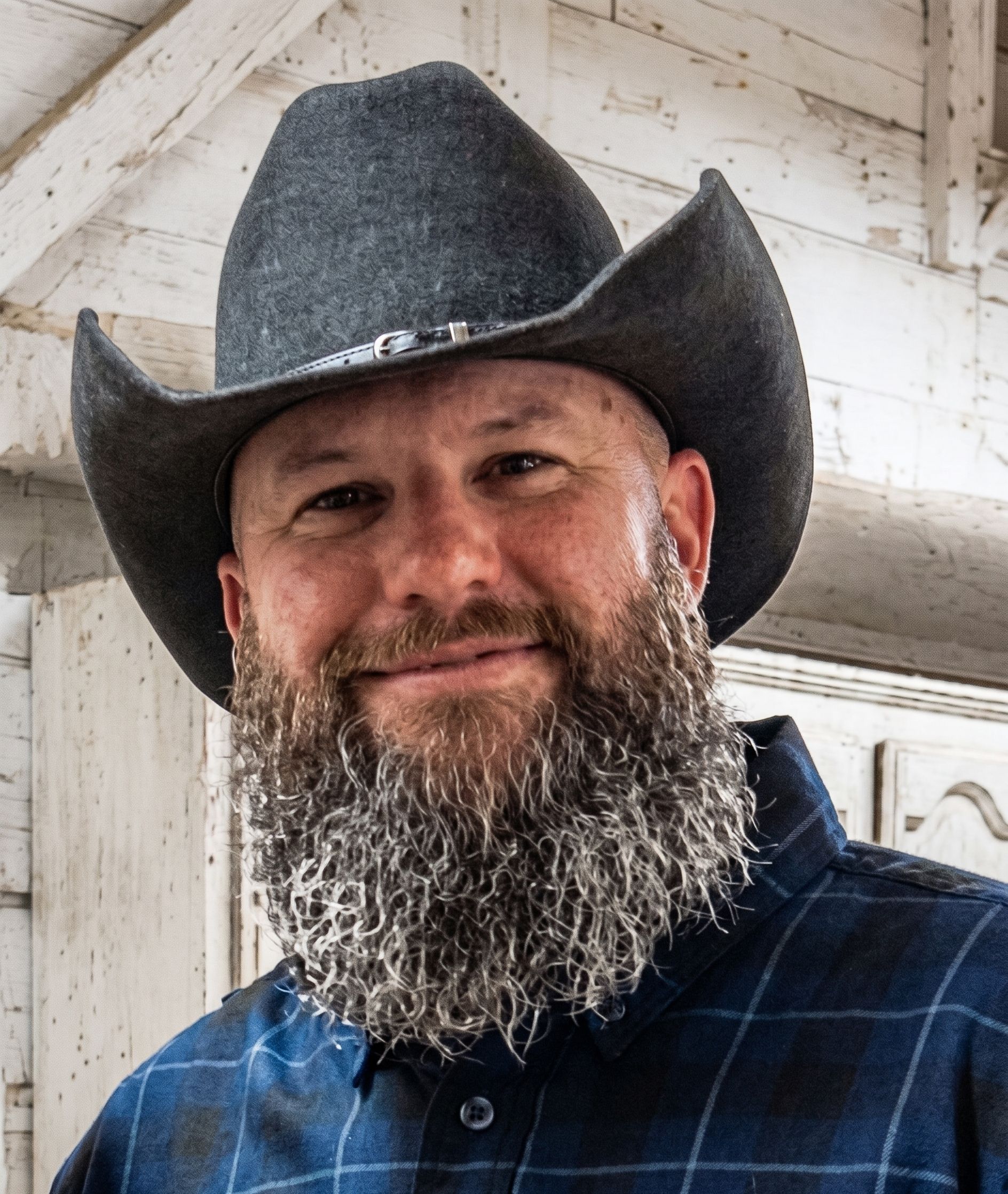 Smiling man in a black cowboy hat and blue plaid shirt standing outdoors by a white wooden wall