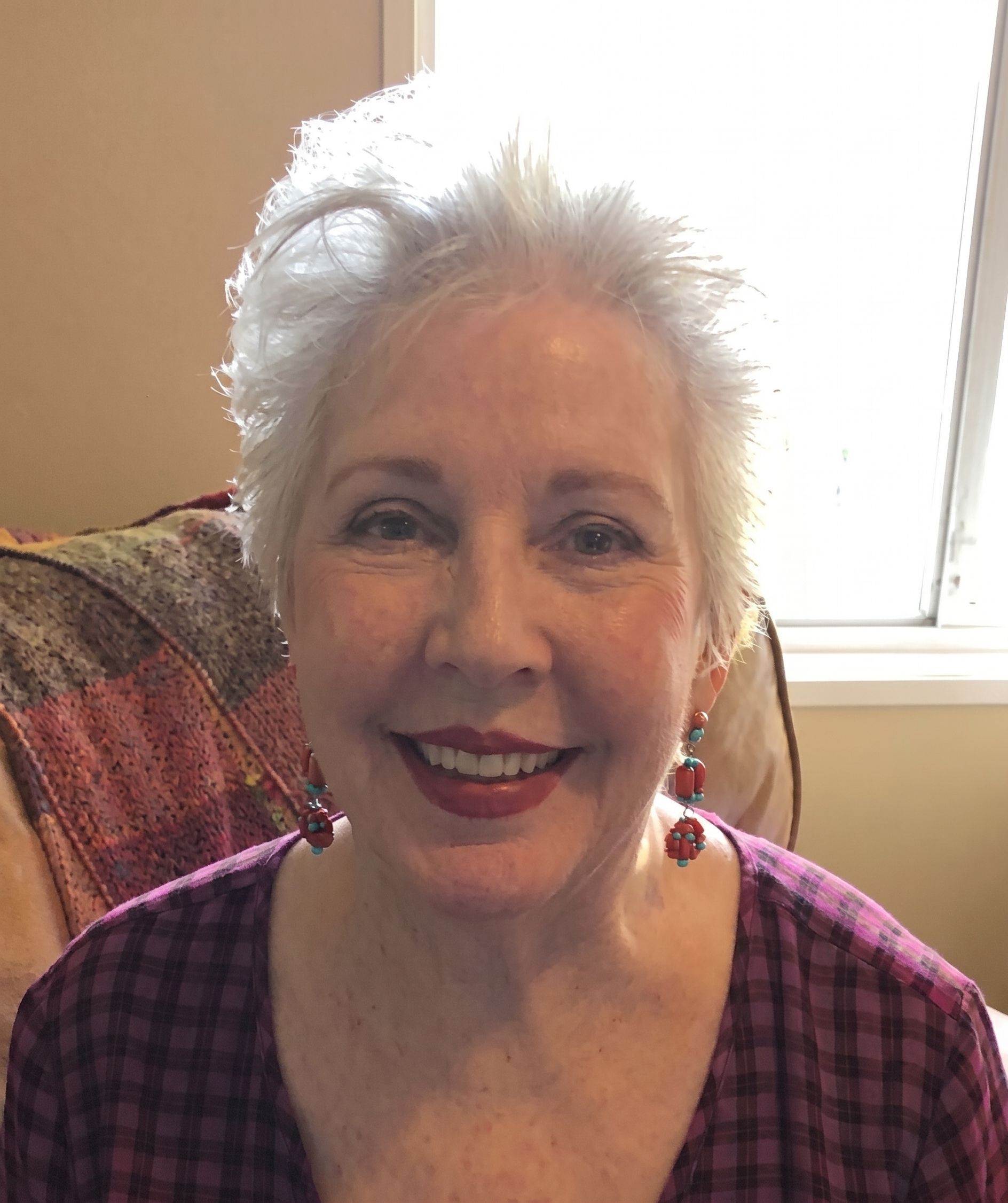 Smiling woman with short white hair in a purple plaid top, sitting by a bright window.