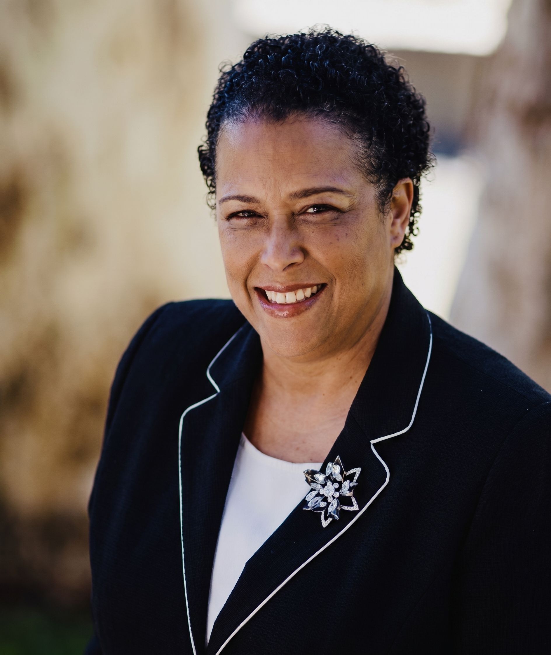 Smiling woman in a black blazer with white trim and a star brooch, standing outdoors.