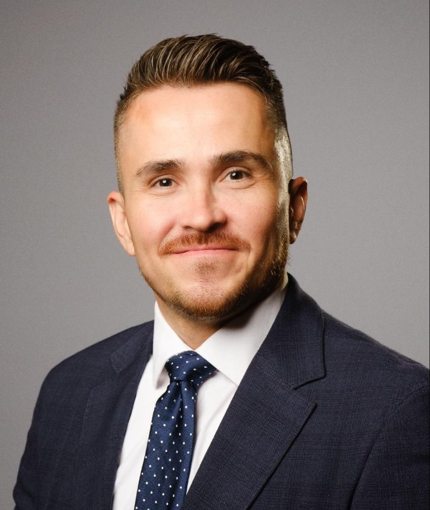 Professional headshot of a man in a navy suit and polka dot tie against a gray background