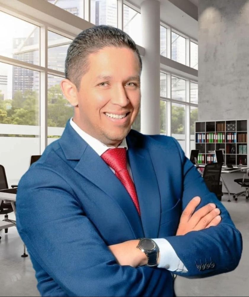 Smiling man in blue suit and red tie standing with arms crossed in a modern office.