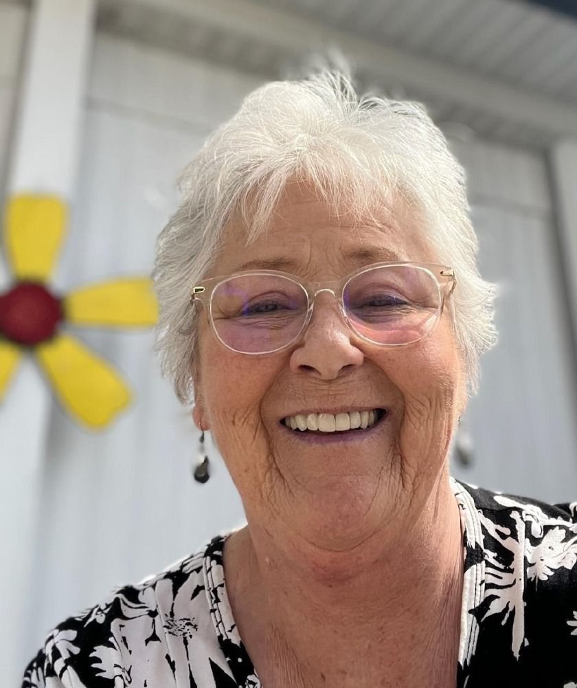 Smiling woman with short white hair and glasses in front of a yellow flower mural.