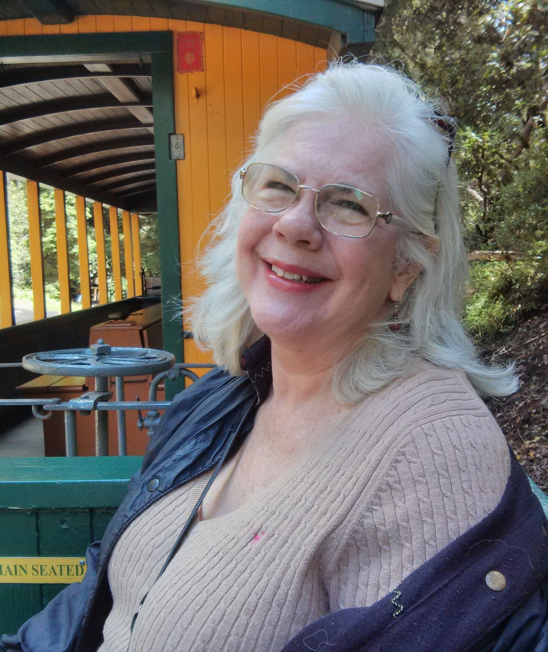 Smiling woman in glasses and beige sweater standing outdoors by a colorful wooden structure